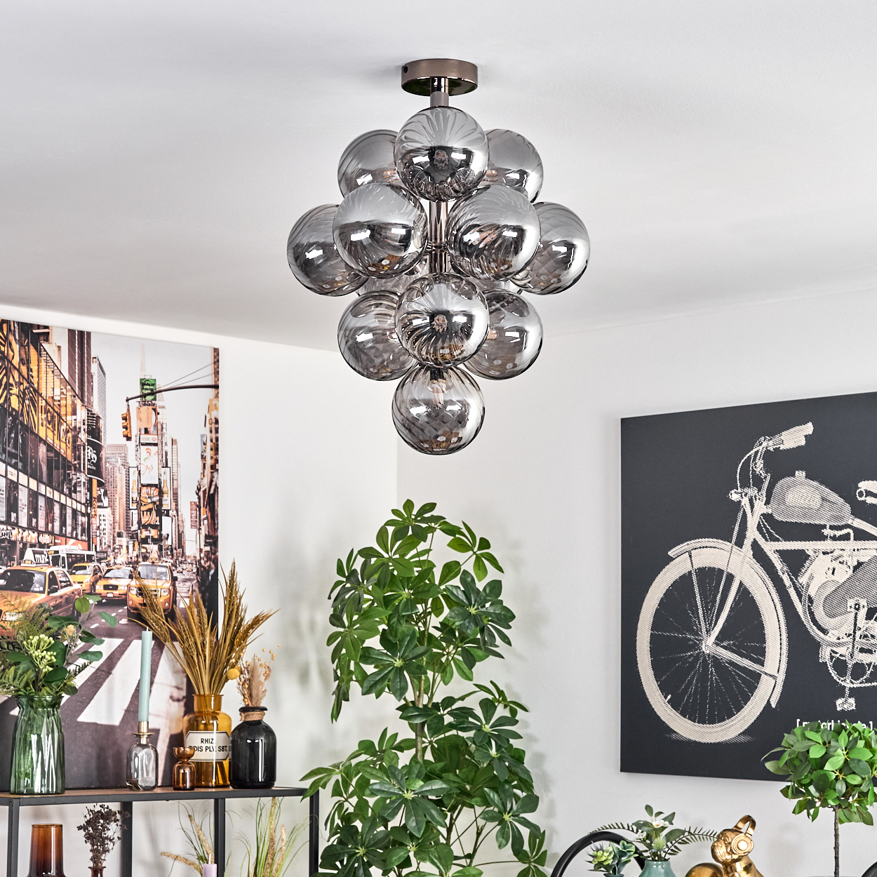 Eine moderne Deckenlampe mit mehreren kugelförmigen Glaskugeln hängt. Ein Stadtbildgemälde und ein Fahrradkunstwerk schmücken die Wände.
