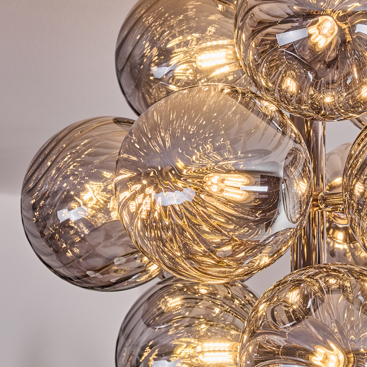 Nahaufnahme eines modernen Kronleuchters. Mehrere rauchfarbene Glaskugeln mit Innenbeleuchtung reflektieren Licht. Chromstützen vor weißem Hintergrund.