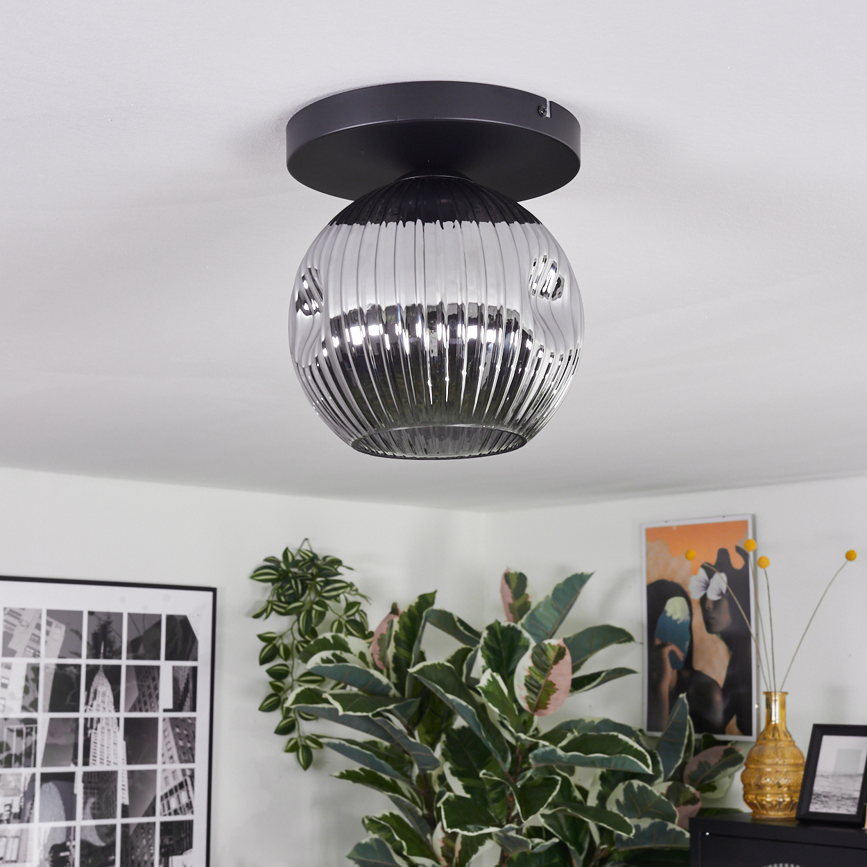 Eine moderne Deckenleuchte mit schwarzem Sockel und geripptem Glasschirm. Pflanzen und Bilder schmücken den Raum und sorgen für Farbe.