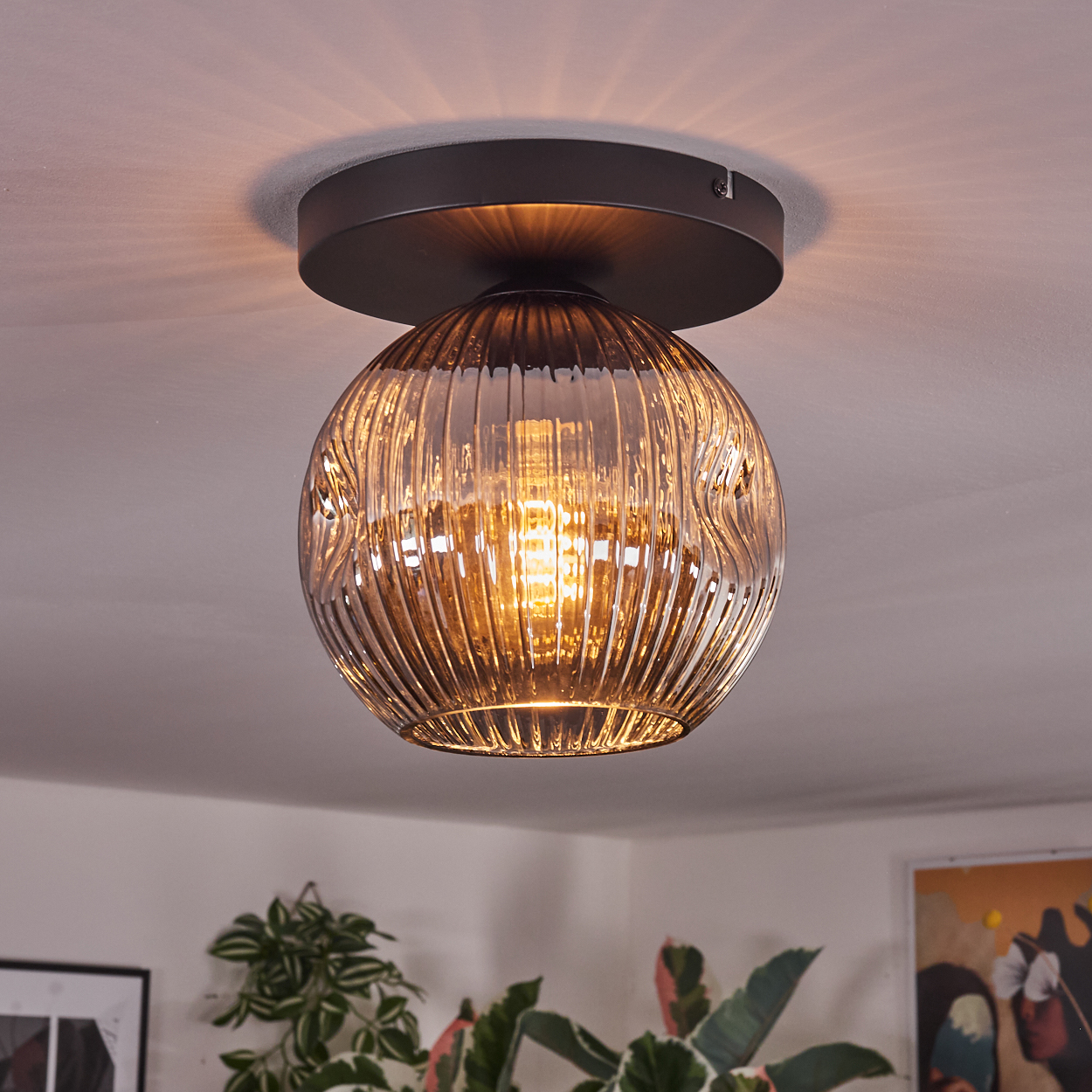 Eine Deckenleuchte mit rundem, strukturiertem Glasschirm. Das Licht scheint durch den Schirm. Pflanzen und Kunst sind im Hintergrund sichtbar.