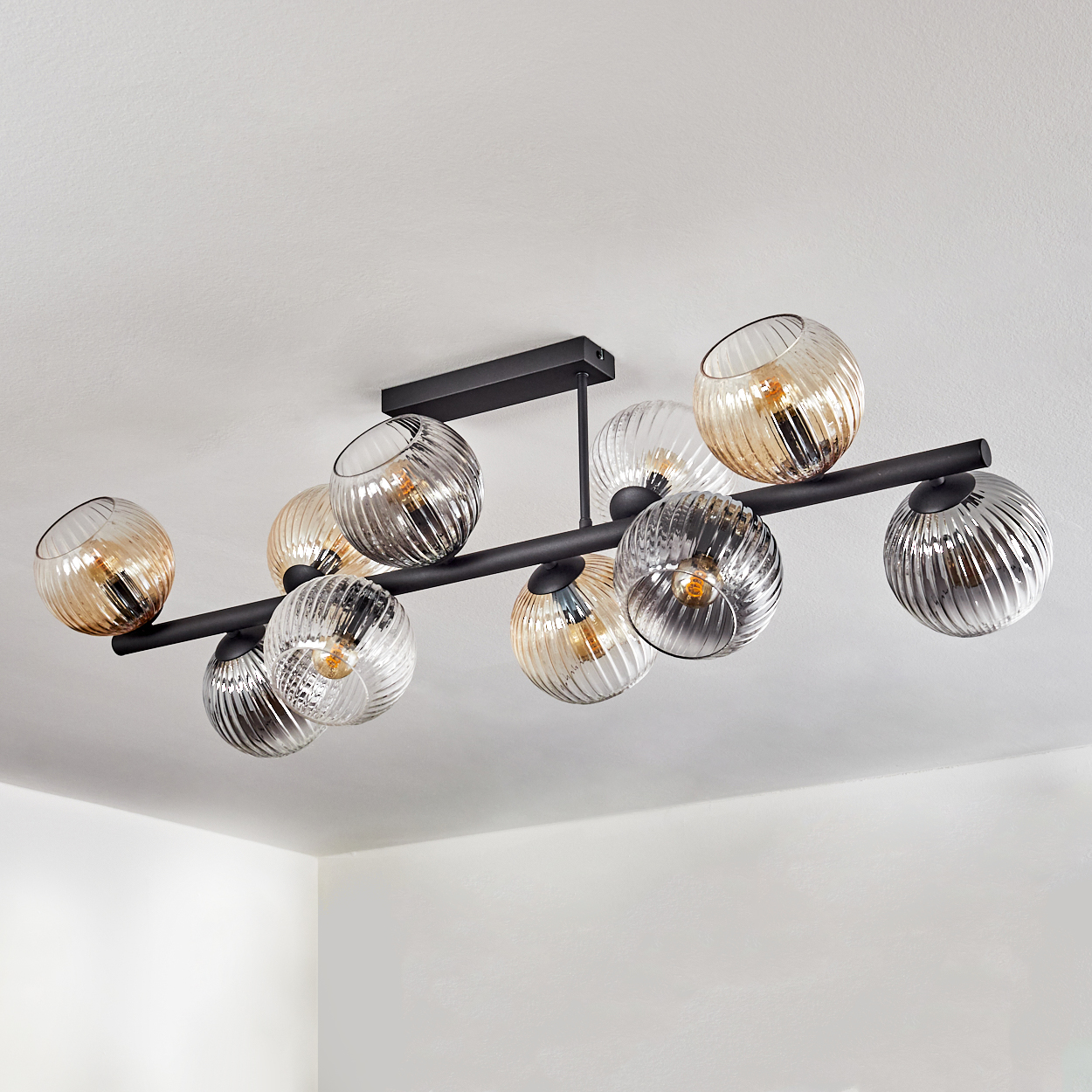 Eine moderne Deckenleuchte mit zehn kugelförmigen Glasschirmen, abwechselnd klar, bernsteinfarben und silber. Schwarzer Metallrahmen. An einer weißen Decke montiert.