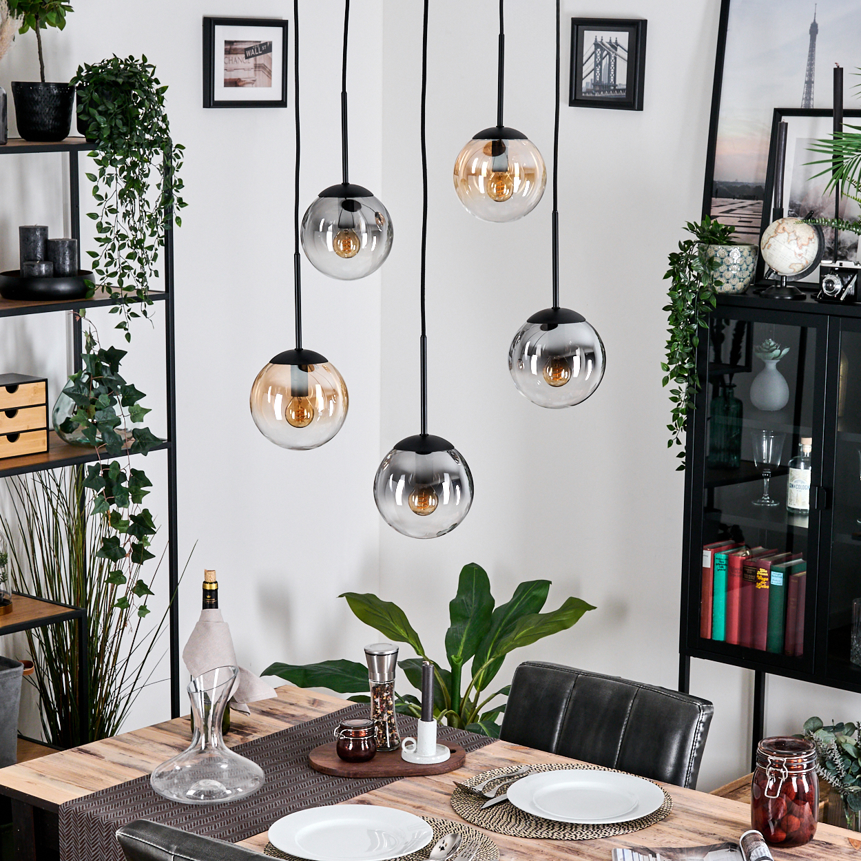 Esszimmer mit Holztisch, Glaskugellampen und schwarzem Schrank. Teller, Gläser und Pflanzen schmücken die Szene.