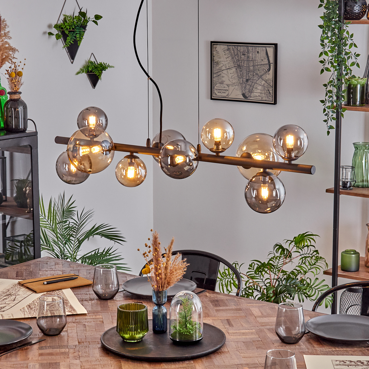 Esszimmer mit Holztisch, modernem Kronleuchter, Pflanzen und Glaswaren. An der Wand hängt eine Karte.