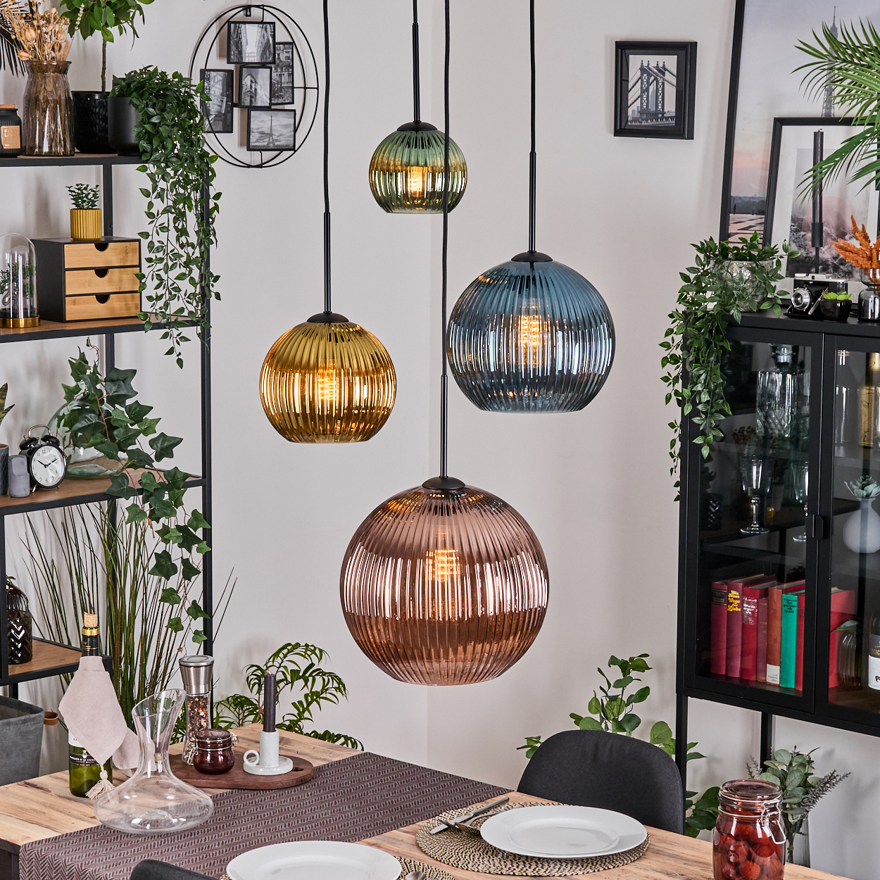 Esszimmer mit vier bunten Hängelampen. Tisch mit Tellern gedeckt. Regale mit Pflanzen, Deko und ein schwarzer Schrank im Hintergrund.