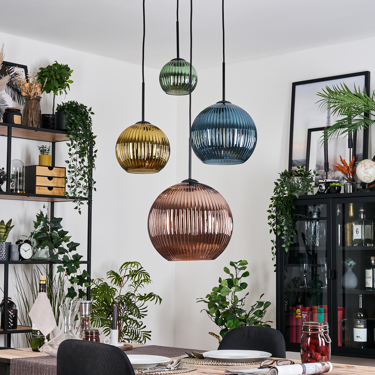 Esszimmer mit vier bunten Glas-Hängelampen. Es gibt Pflanzen, einen Tisch und einen schwarzen Schrank mit Bildern.