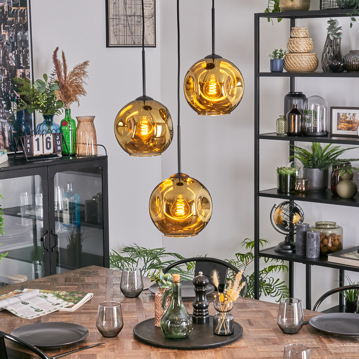Esszimmer mit drei goldenen Pendelleuchten. Tisch gedeckt mit Tellern, Gläsern und Dekor. Regale mit Pflanzen und Dekor.