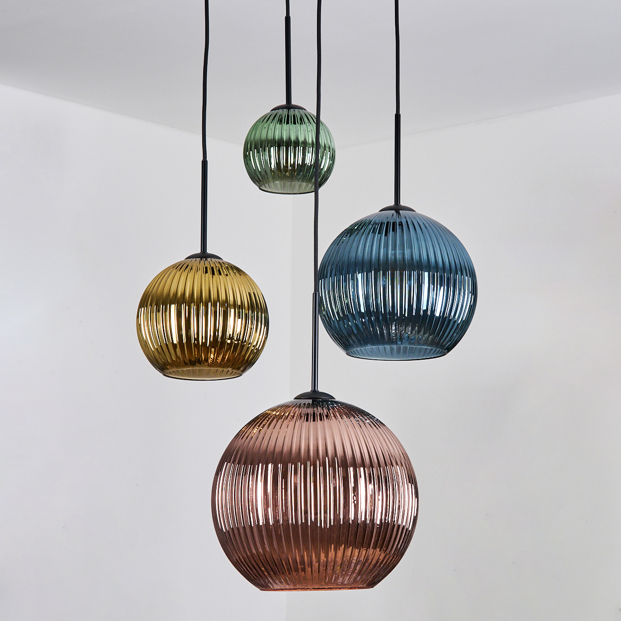 Vier bunte, gerippte Glas-Pendelleuchten hängen an schwarzen Kabeln vor einer weißen Wand. Die Farben sind Gold, Grün, Blau und Rosé.