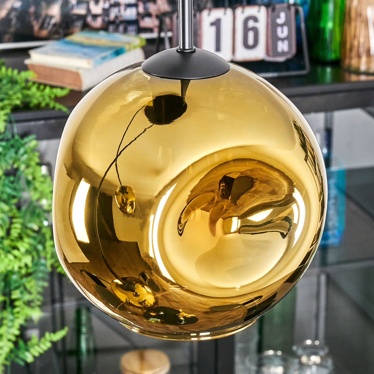 Eine goldene, kugelförmige Hängelampe hängt. Sie reflektiert den Raum und die umliegenden Objekte. Grüne Pflanzen und Bücher sind im Hintergrund sichtbar.
