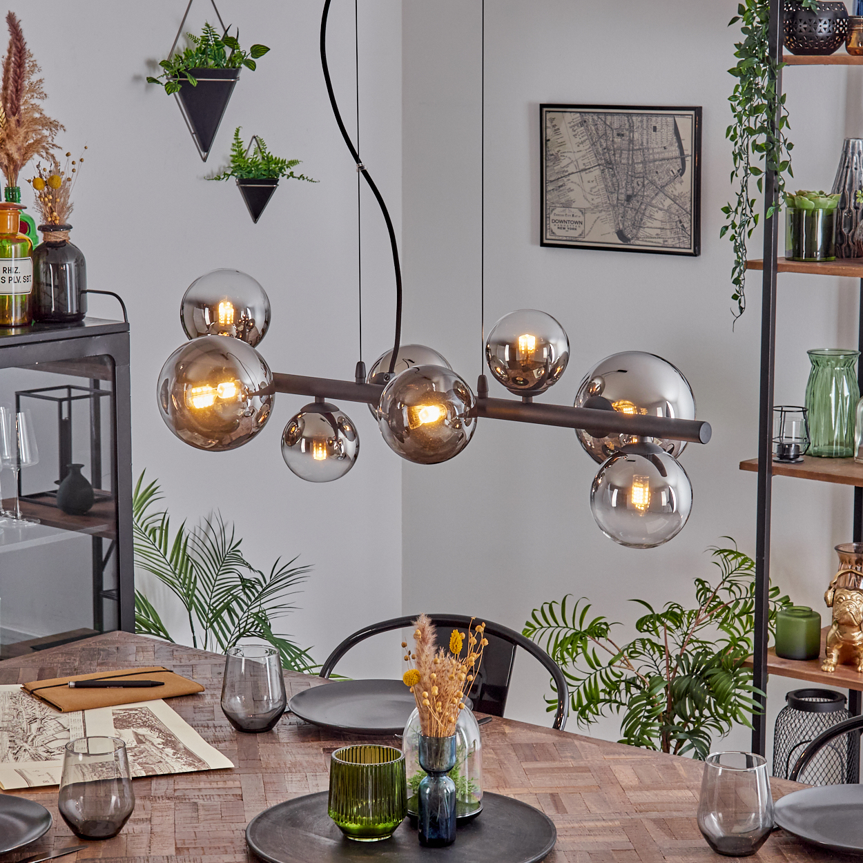 Ein Esszimmer mit Holztisch, modernem Kronleuchter, Gläsern und Zierpflanzen. Im Hintergrund ein schwarzes Metallregal.