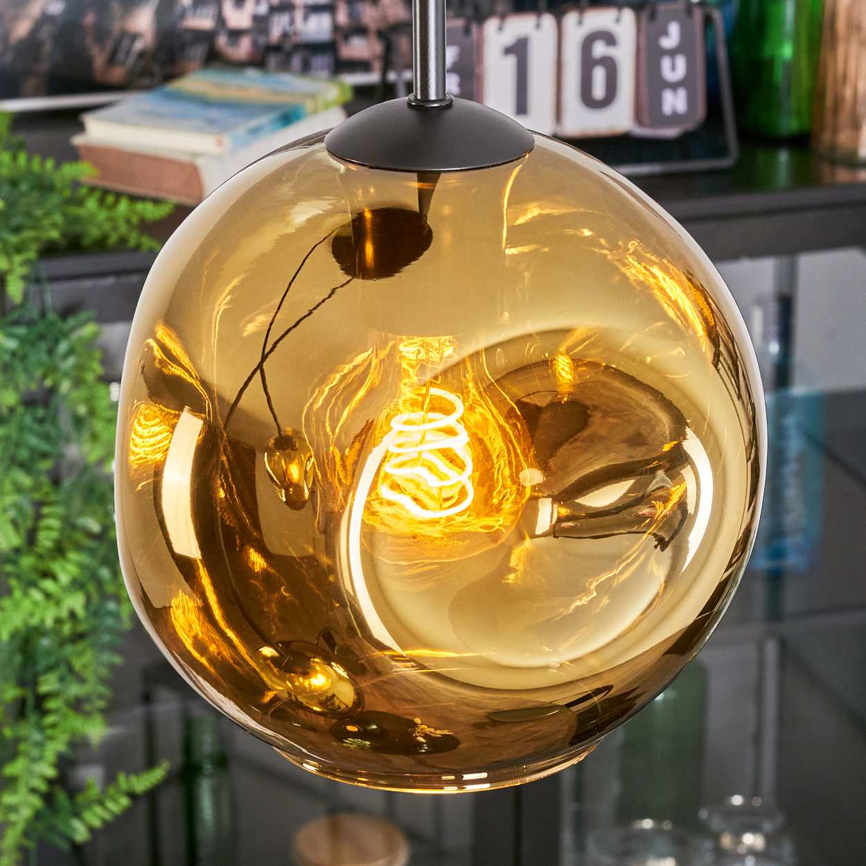 Eine goldene Kugel-Pendelleuchte mit einer Wendelbirne im Inneren, die von der Decke hängt. Im Hintergrund sind ein Kalender und Grün zu sehen.