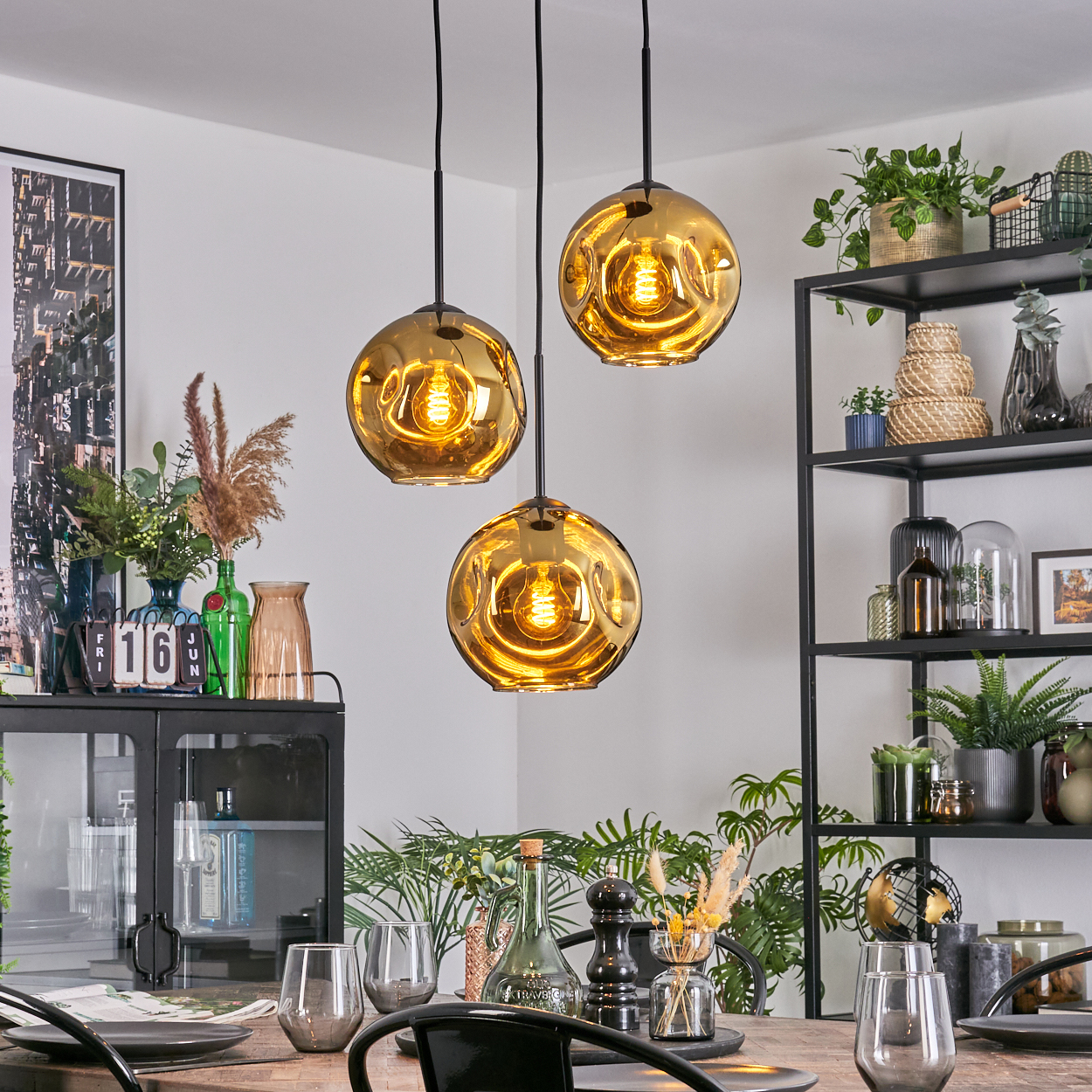 Esszimmer mit drei goldenen Pendelleuchten, gedecktem Tisch und Pflanzen auf Regalen.