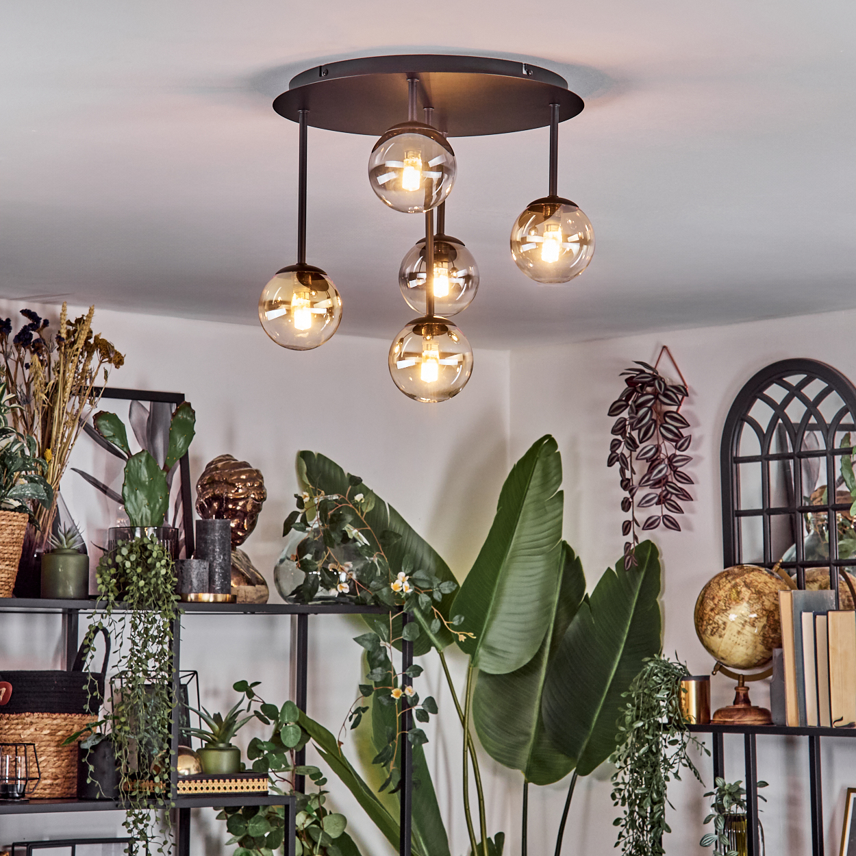 Ein modernes Wohnzimmer mit einem schwarzen Metallregal voller Pflanzen und Dekoration. Über dem Raum hängt eine runde Deckenlampe mit Glaskugeln.