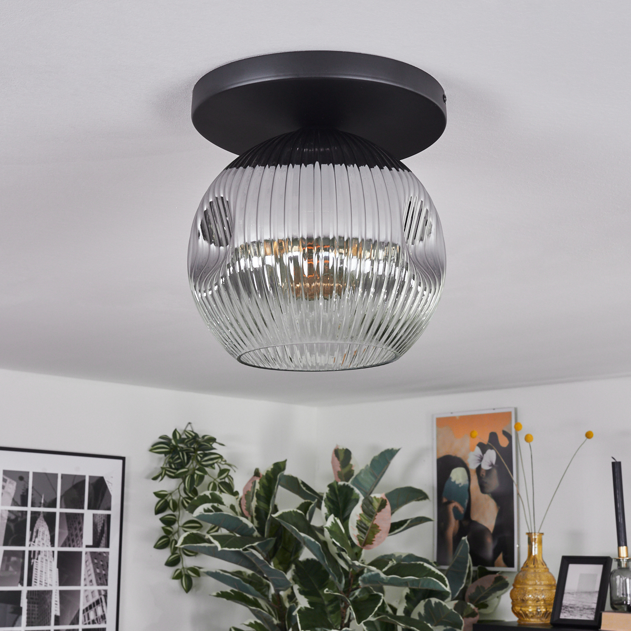 Eine moderne Deckenleuchte mit schwarzem Sockel und geripptem Glasschirm. Pflanzen und Kunstwerke sind im Hintergrund zu sehen.