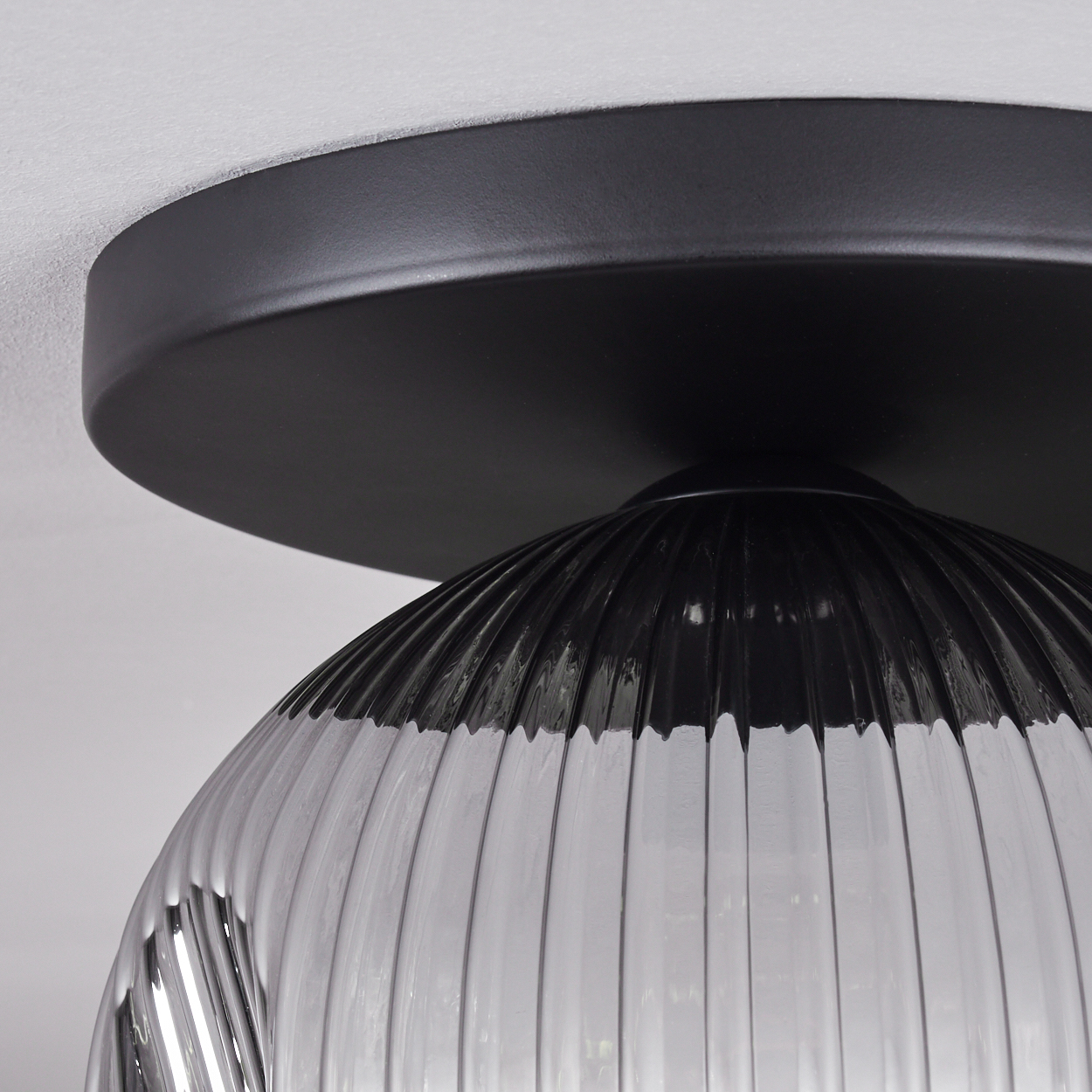 Nahaufnahme einer schwarzen Deckenlampe. Sie hat einen runden, schwarzen Sockel und einen gerippten, klaren Glasschirm. Der Hintergrund ist weiß.