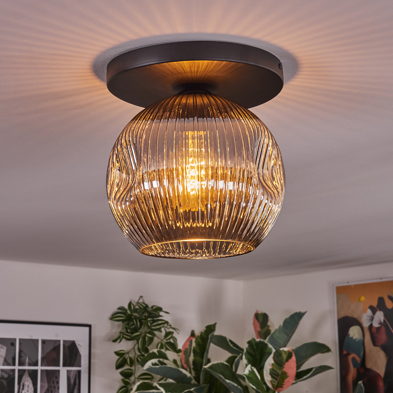 Nahaufnahme einer Deckenleuchte. Sie hat einen runden, geriffelten Glasschirm und einen dunklen Metallsockel. Pflanzen und Kunst sind im Hintergrund.