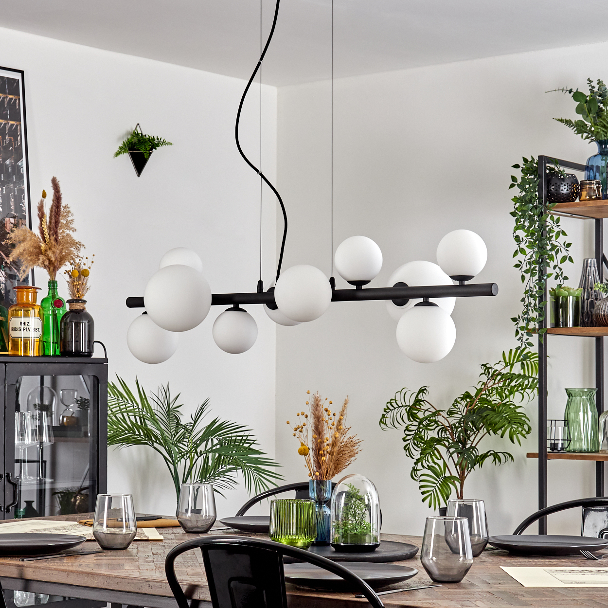 Esszimmer mit Holztisch, schwarzen Stühlen und einer schwarzen Leuchte mit weißen Kugeln. Dekorationen umfassen Pflanzen und Glaswaren.