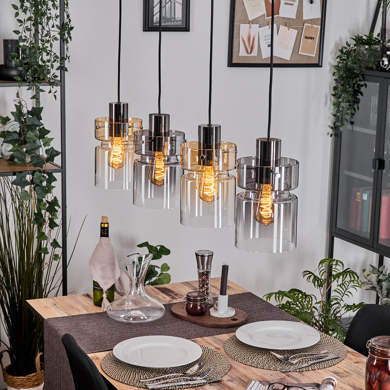 Vier moderne Pendelleuchten hängen über einem Holztisch, der für eine Mahlzeit gedeckt ist. Der Hintergrund umfasst Pflanzen, Regale und einen Schrank.
