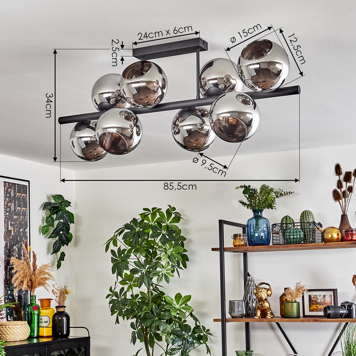 Eine moderne Deckenlampe mit silbernen Kugeln, im Hintergrund ein Wohnzimmer mit Pflanzen und Regalen mit Dekorationsartikeln.