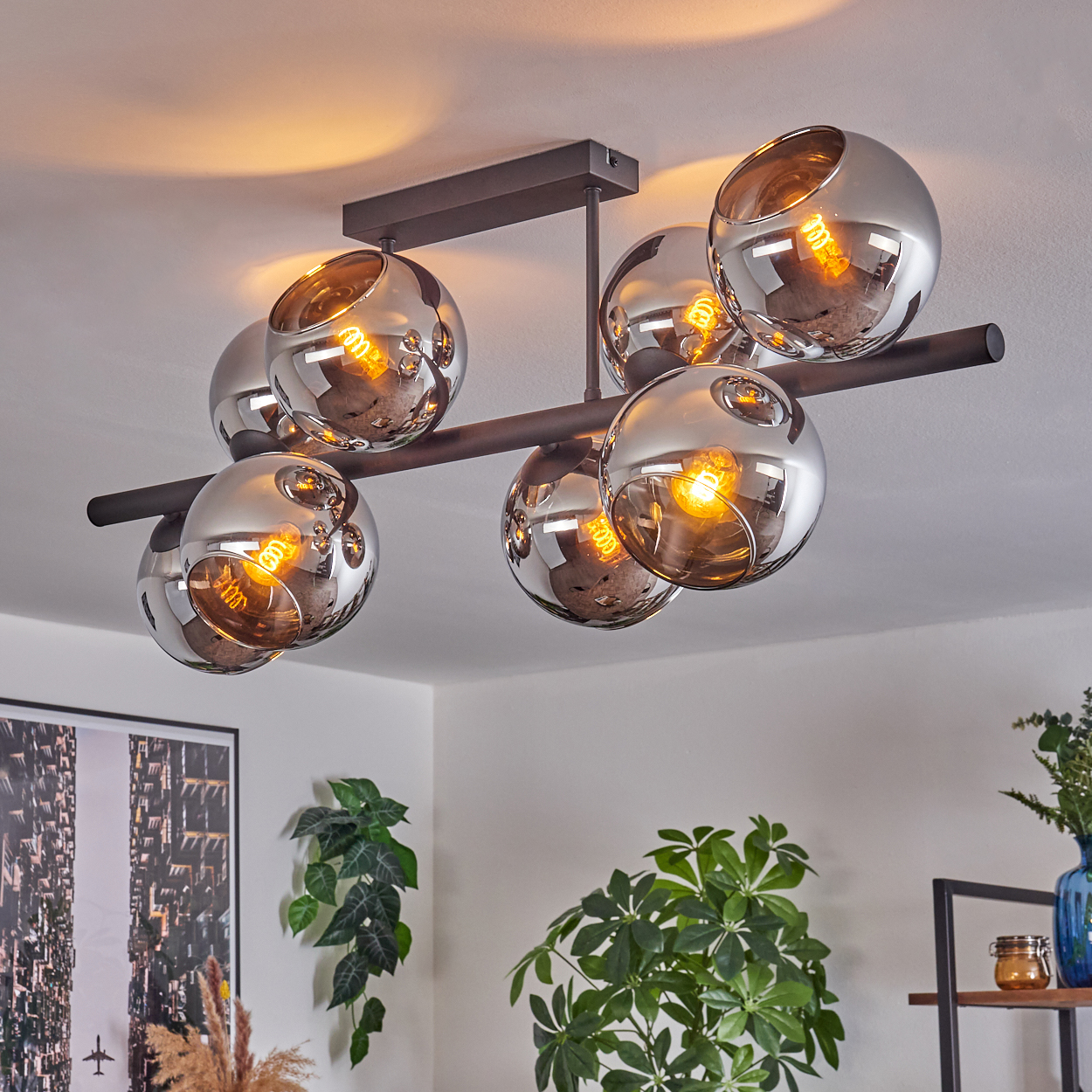 Eine moderne Deckenlampe mit sechs kugelförmigen, verspiegelten Glasschirmen, jede mit einer warm leuchtenden Glühbirne. Der Raum hat Pflanzen und ein Stadtbildgemälde.
