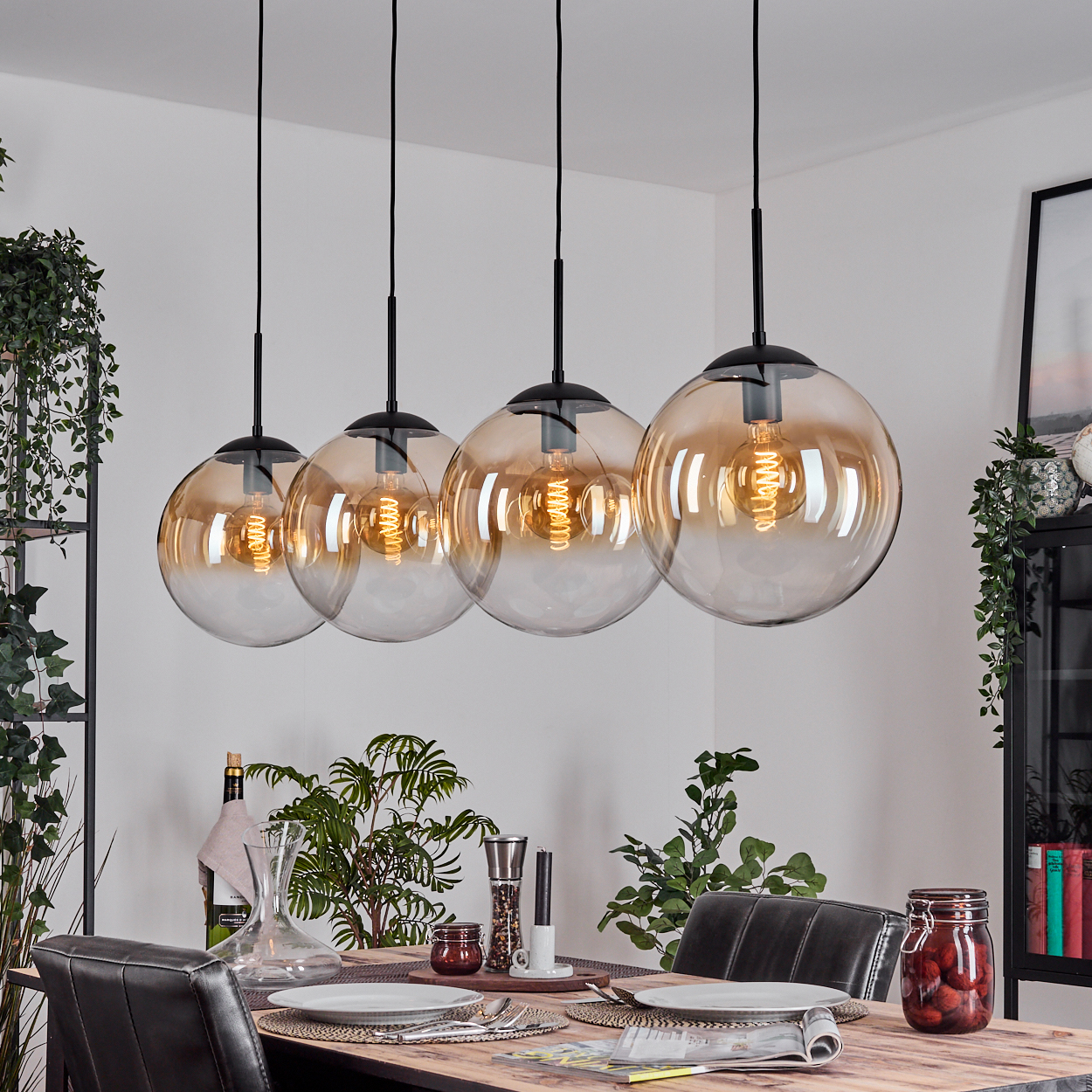 Vier Pendelleuchten aus bernsteinfarbenem Glas hängen über einem Holzesstisch, der für eine Mahlzeit gedeckt ist. Pflanzen und Dekoration umgeben den Tisch.