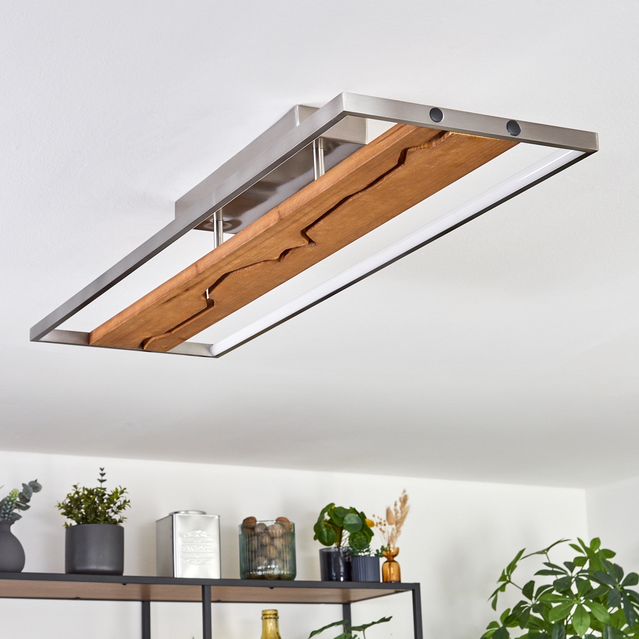 Eine moderne Deckenleuchte mit Holzmitte und Metallrahmen. Darunter ein Regal mit Pflanzen und Dekoration vor weißer Wand.