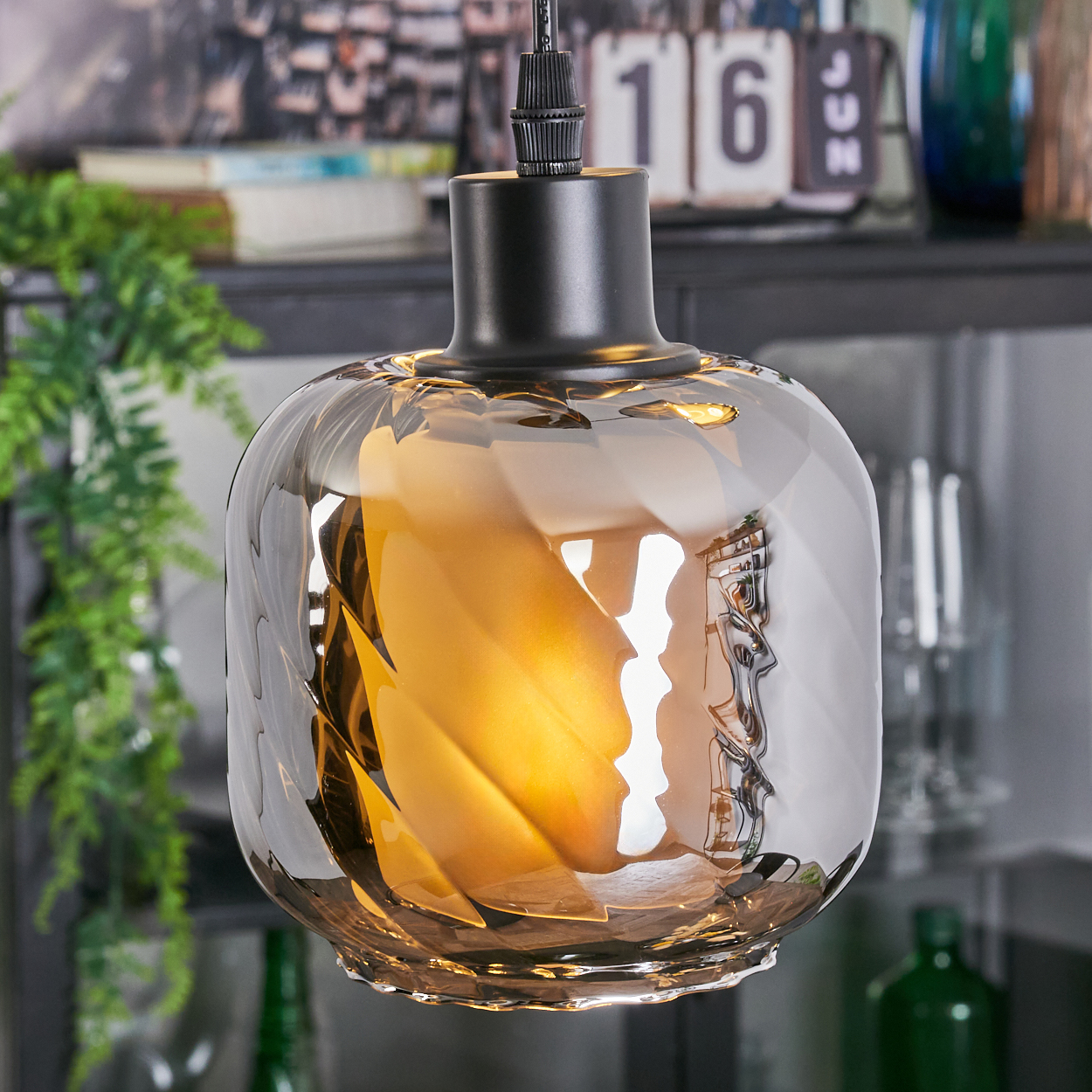 Eine dekorative Glas-Pendelleuchte mit strukturierter Oberfläche, die warmes Licht ausstrahlt. Hintergrund: Regal, Kalender und Grün.