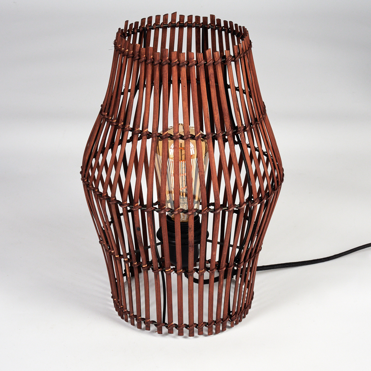Eine braune Bambuslampe mit einer Glühbirne im Inneren, auf weißem Hintergrund. Ein schwarzes Kabel ist sichtbar.