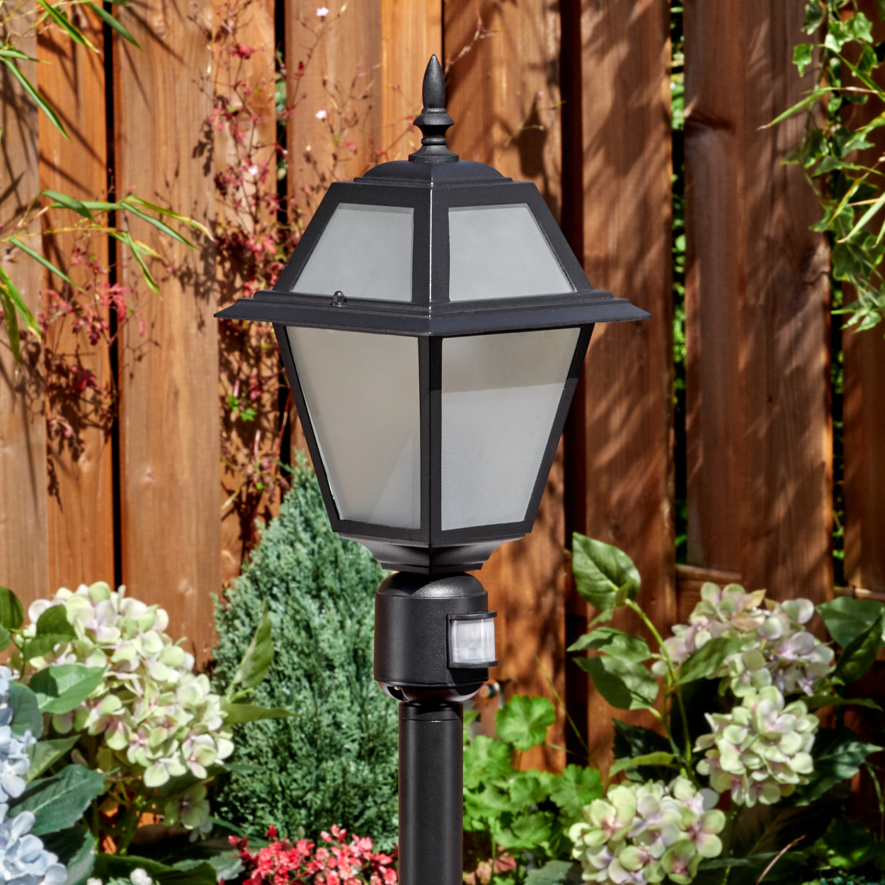 Schwarze Außenlampe mit Milchglas. Sie steht auf einer Stange in einem Garten. Holzzaun und Pflanzen sind im Hintergrund.