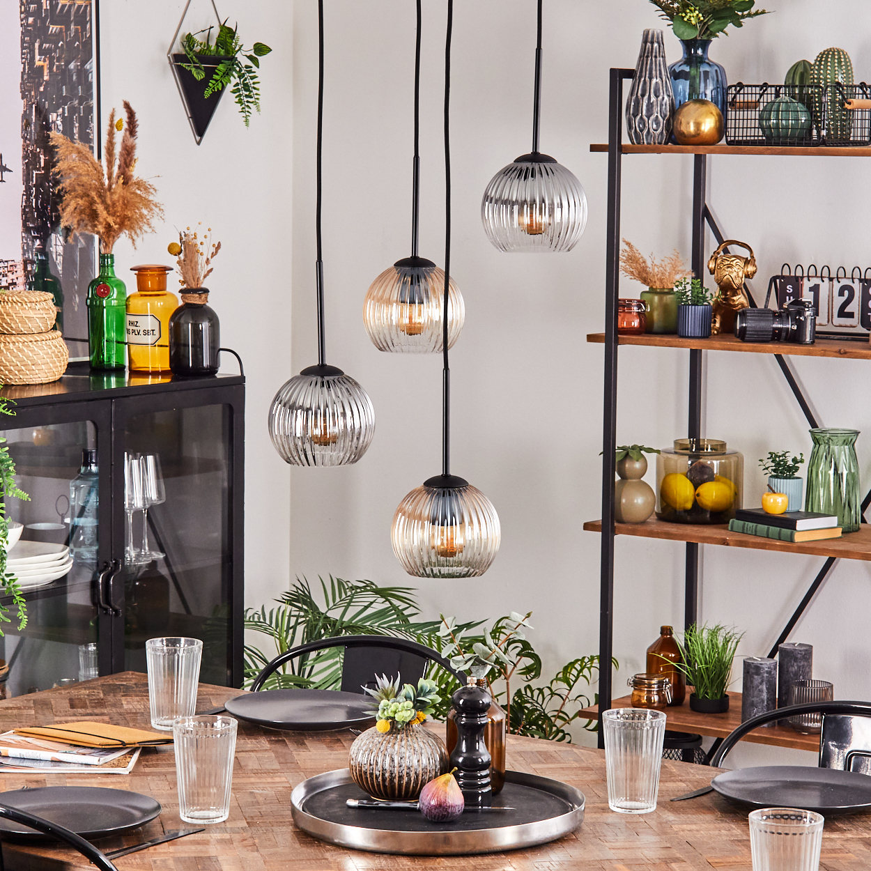 Esszimmer mit Holztisch, Glaslampen und dekorativen Gegenständen. Teller, Gläser und ein Tablett mit Vase und Feige stehen auf dem Tisch.