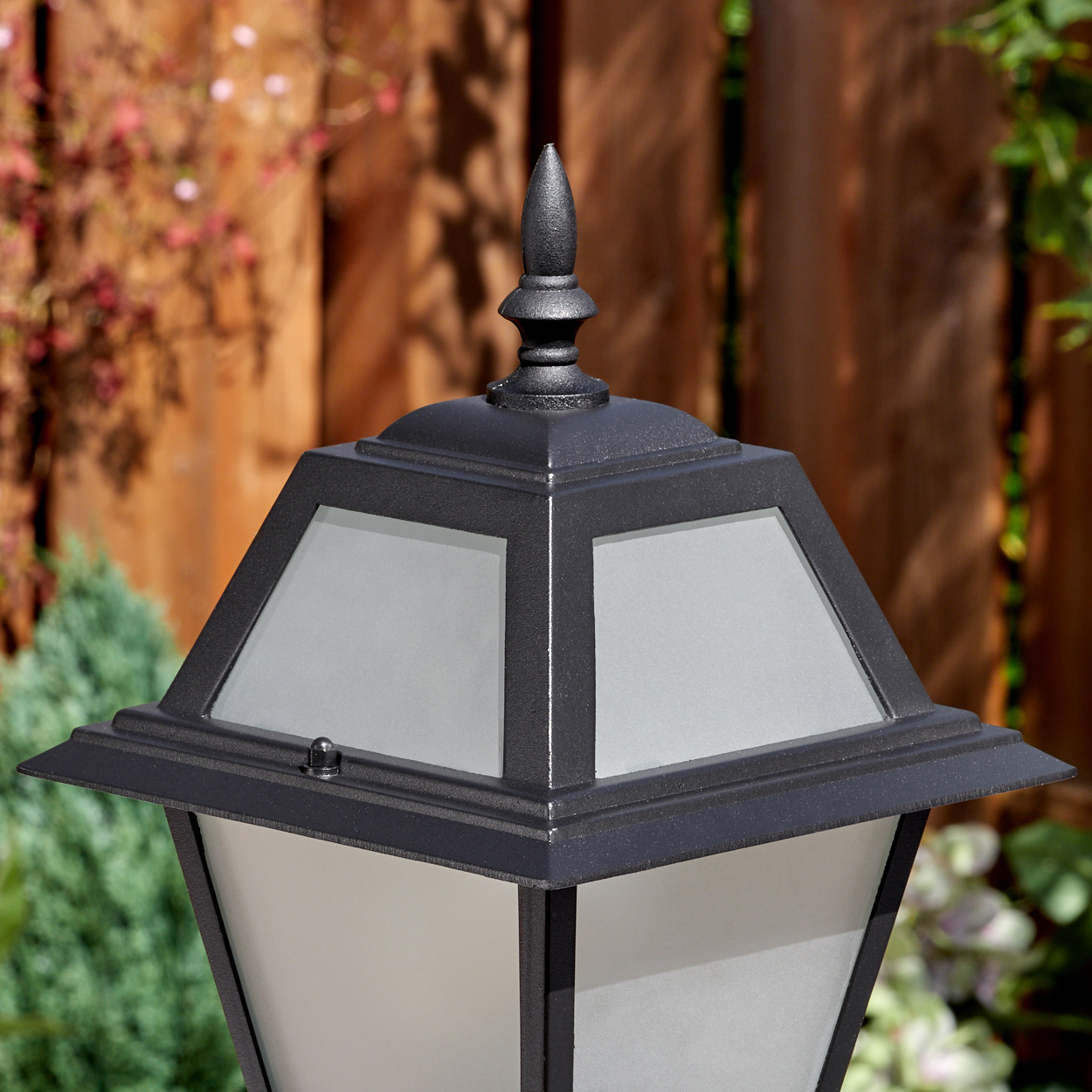 Nahaufnahme einer schwarzen Außenlampe mit Milchglas. Ein Holzzaun und Grünzeug sind im verschwommenen Hintergrund.