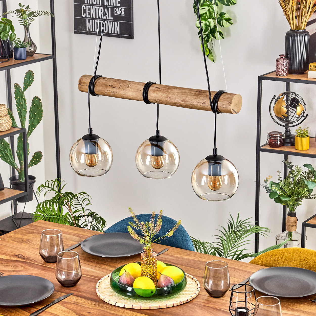 Esszimmer mit Holztisch, Glaslampen, Pflanzen, Tellern, Gläsern und einer Obstschale. Ein Schild hängt an der Wand.