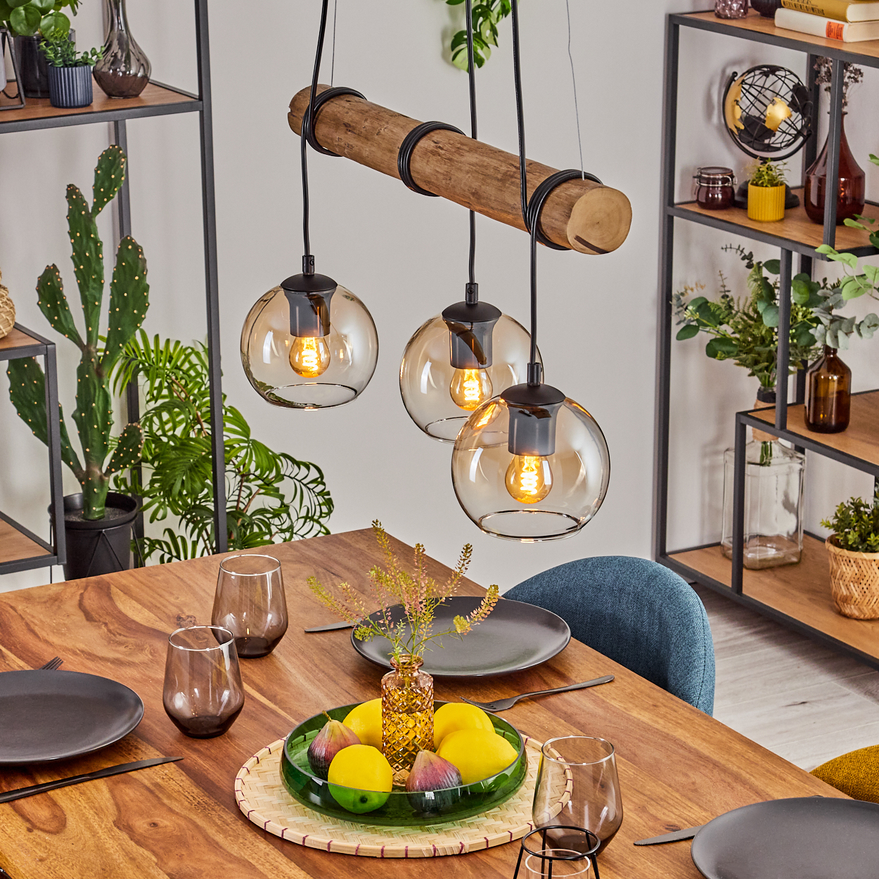 Ein Esstisch mit Obst, Gläsern und Tellern. Oben hängen drei leuchtende Pendelleuchten von einem Holzbalken.