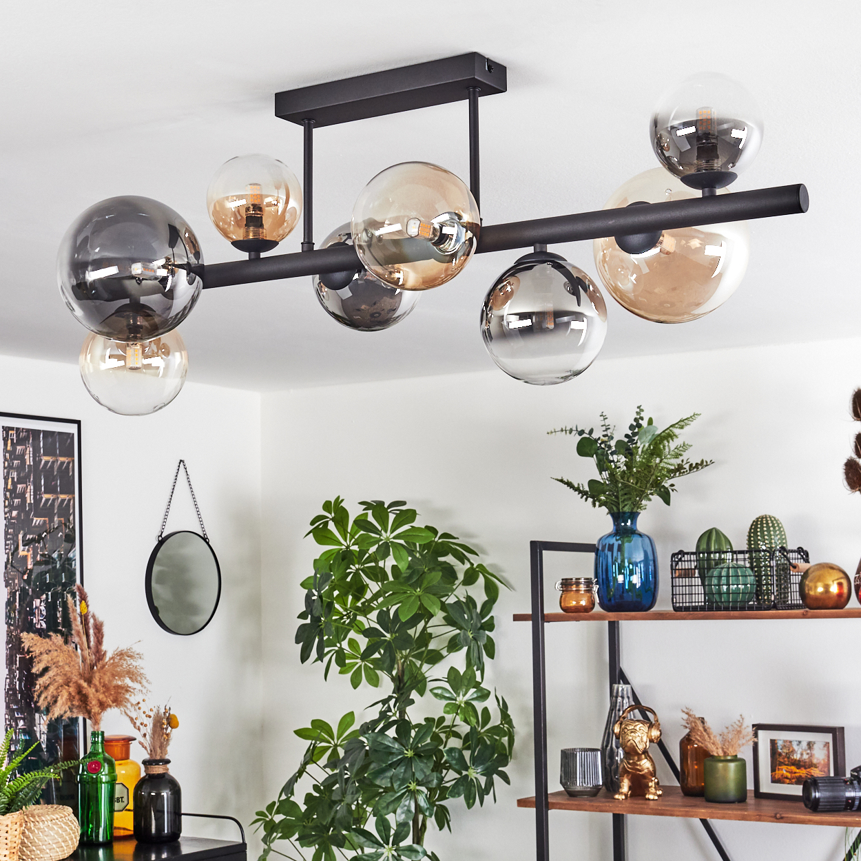 Eine moderne schwarze Hängelampe mit Glaskugeln hängt. Darunter Pflanzen, ein Spiegel und Regale mit Dekoration. Neutrale Wand, gemütliches Interieur.