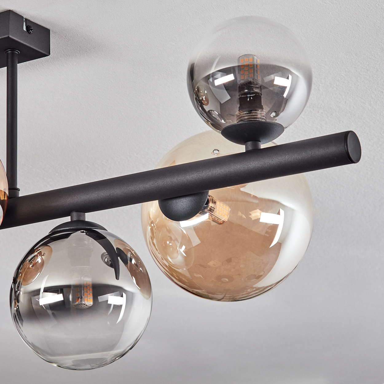 Eine moderne Deckenleuchte mit Glaskugelschirmen, schwarzem Metallrahmen und sichtbaren Glühbirnen vor weißer Decke.