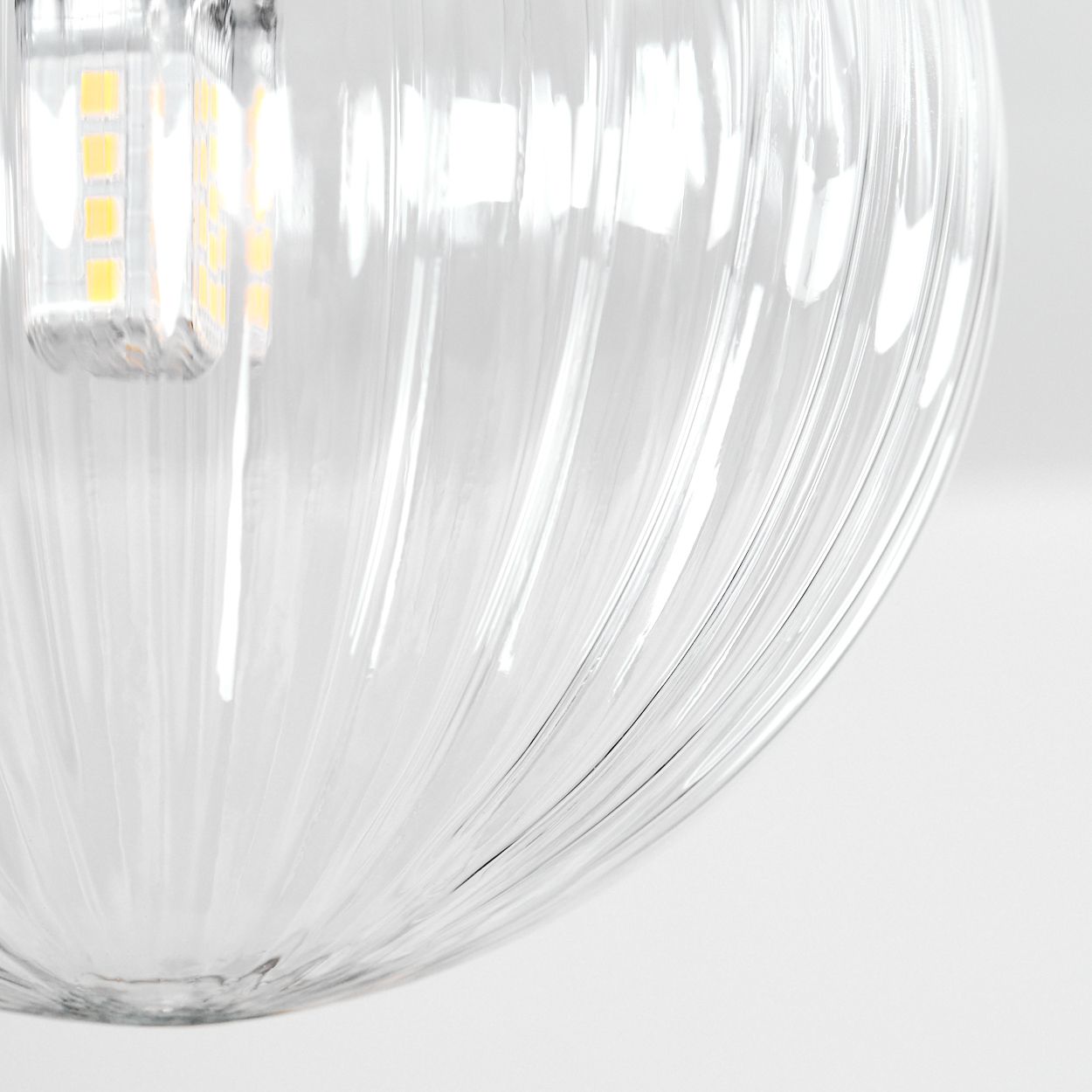 Nahaufnahme eines Lampenschirms aus klarem Glas mit vertikalen Rippen. Eine helle LED-Glühbirne ist oben links vor weißem Hintergrund sichtbar.