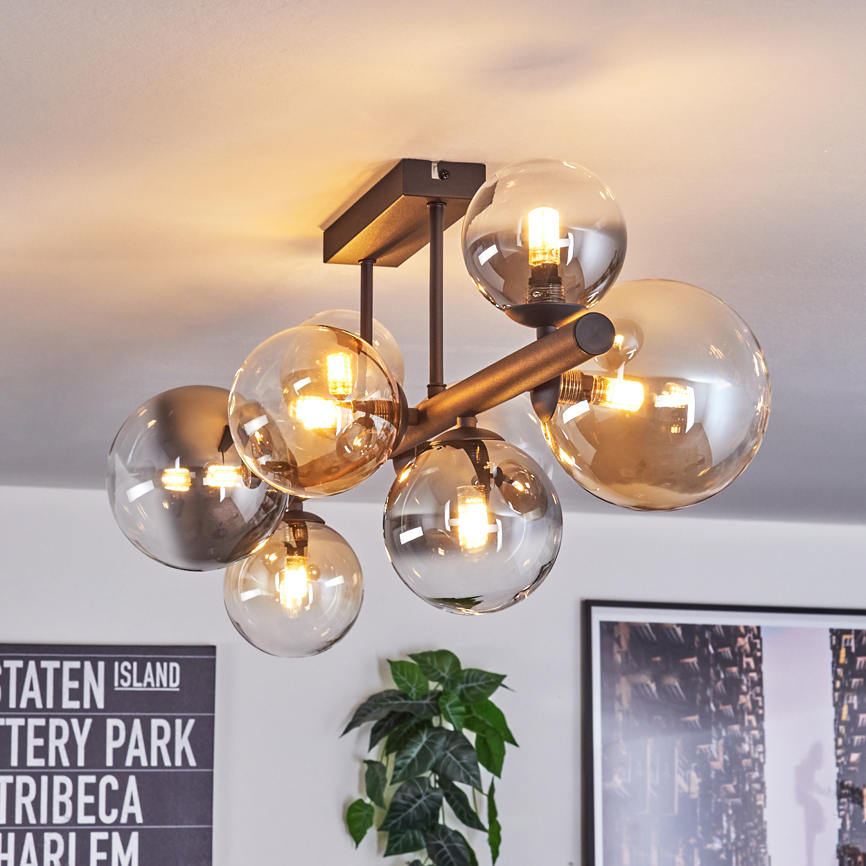 Eine moderne Deckenleuchte mit Glaskugeln. Sie hat einen dunklen Metallrahmen und warm getönte Glühbirnen. Im Hintergrund sind eine Pflanze und Kunstwerke.