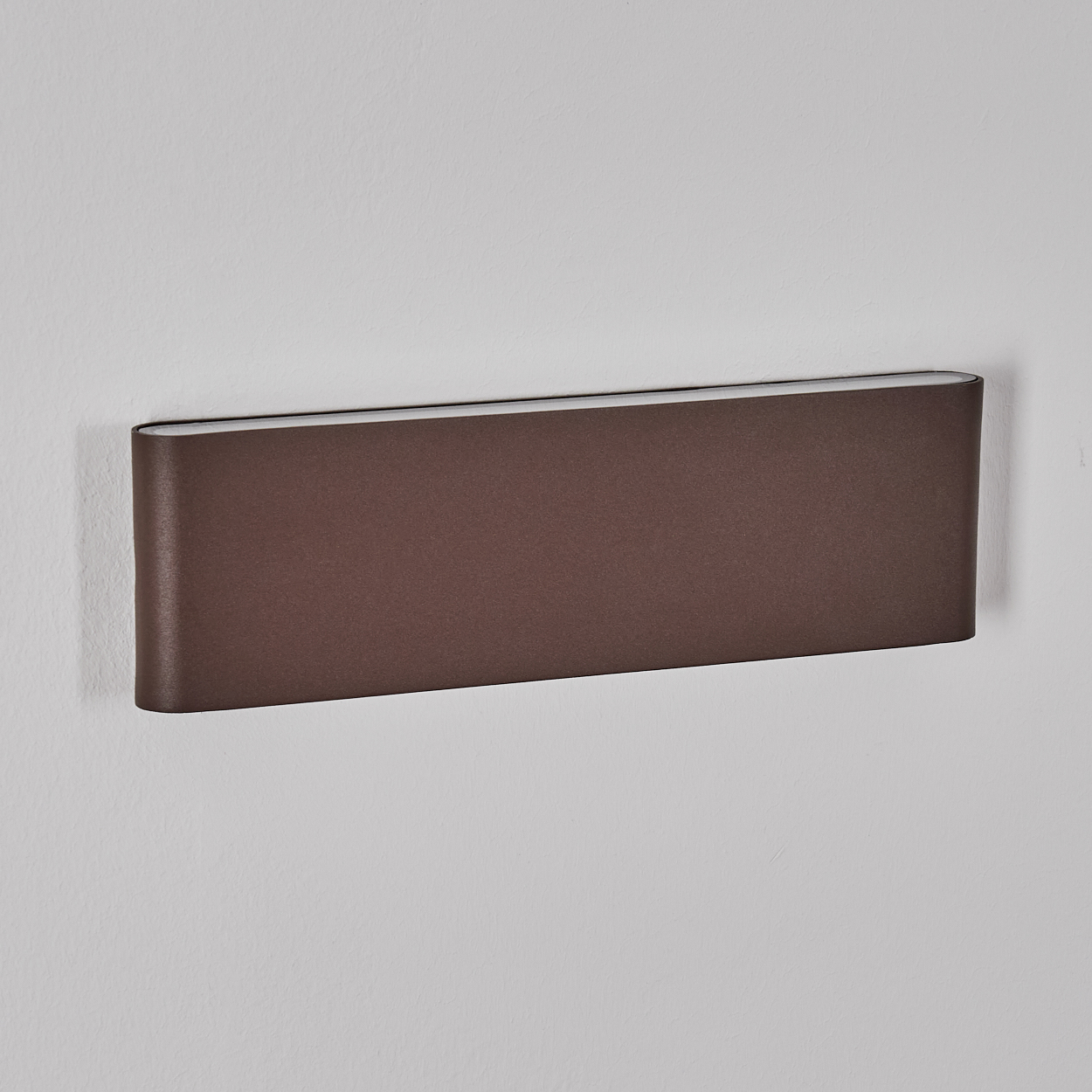 Eine braune, rechteckige Wandlampe ist an einer weißen Wand montiert. Die Lampe hat abgerundete Kanten und eine hellere Innenseite.