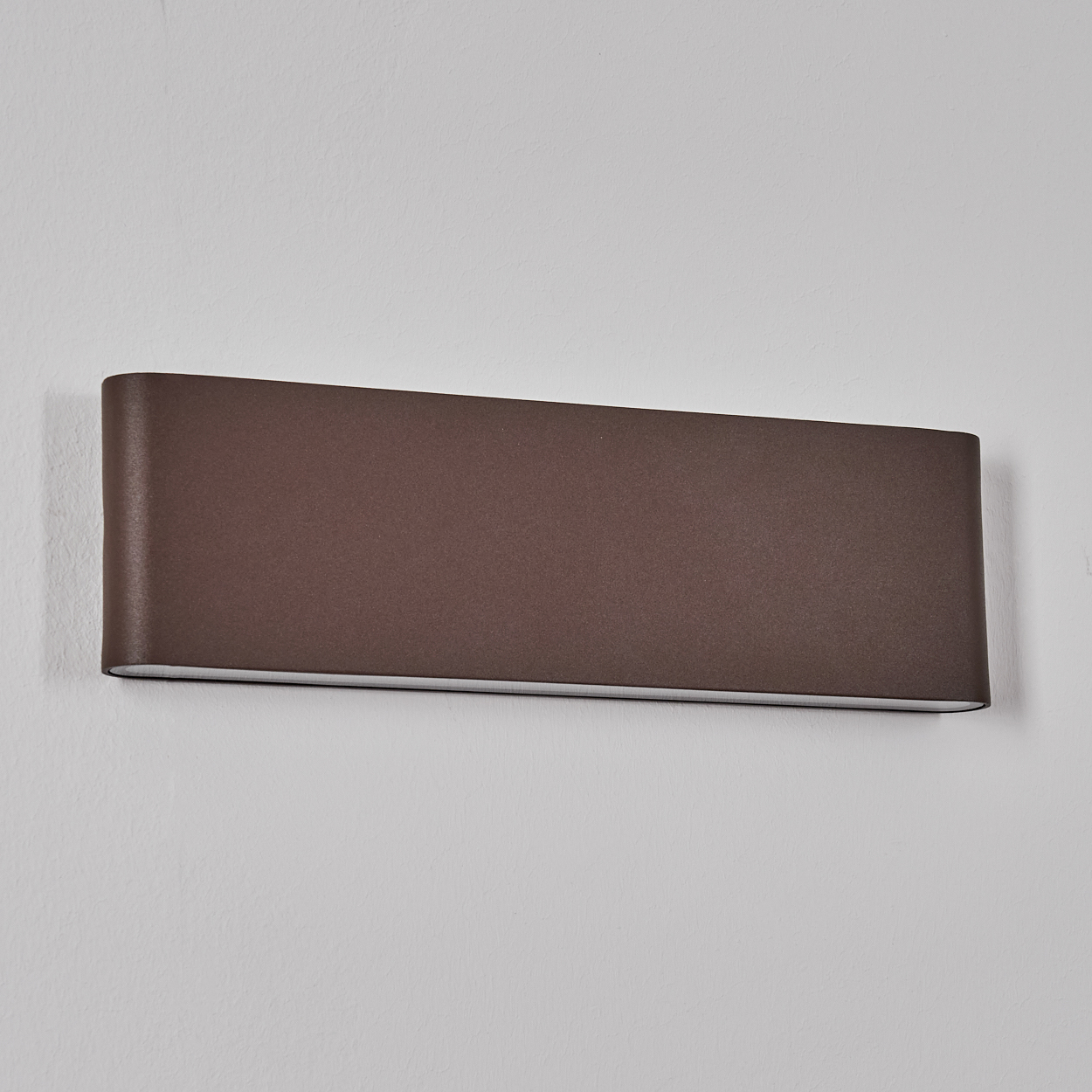 Eine braune, rechteckige Wandleuchte ist an einer weißen Wand befestigt. Die Lampe hat eine gebogene Form und ist horizontal angeordnet.