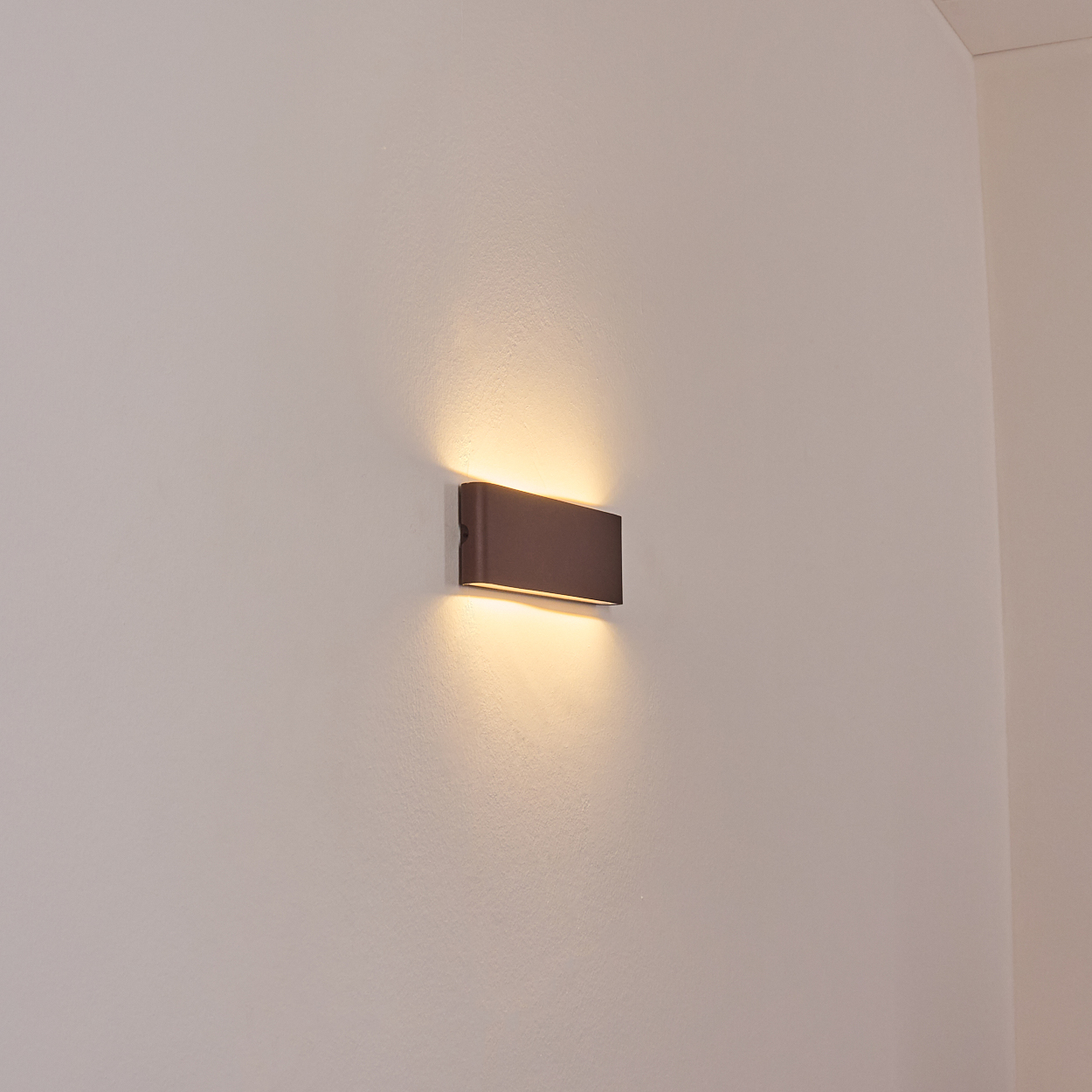 Eine rechteckige, braune Wandleuchte strahlt warmes Licht aus. Sie ist an einer weißen Wand in der Ecke eines Raumes montiert.