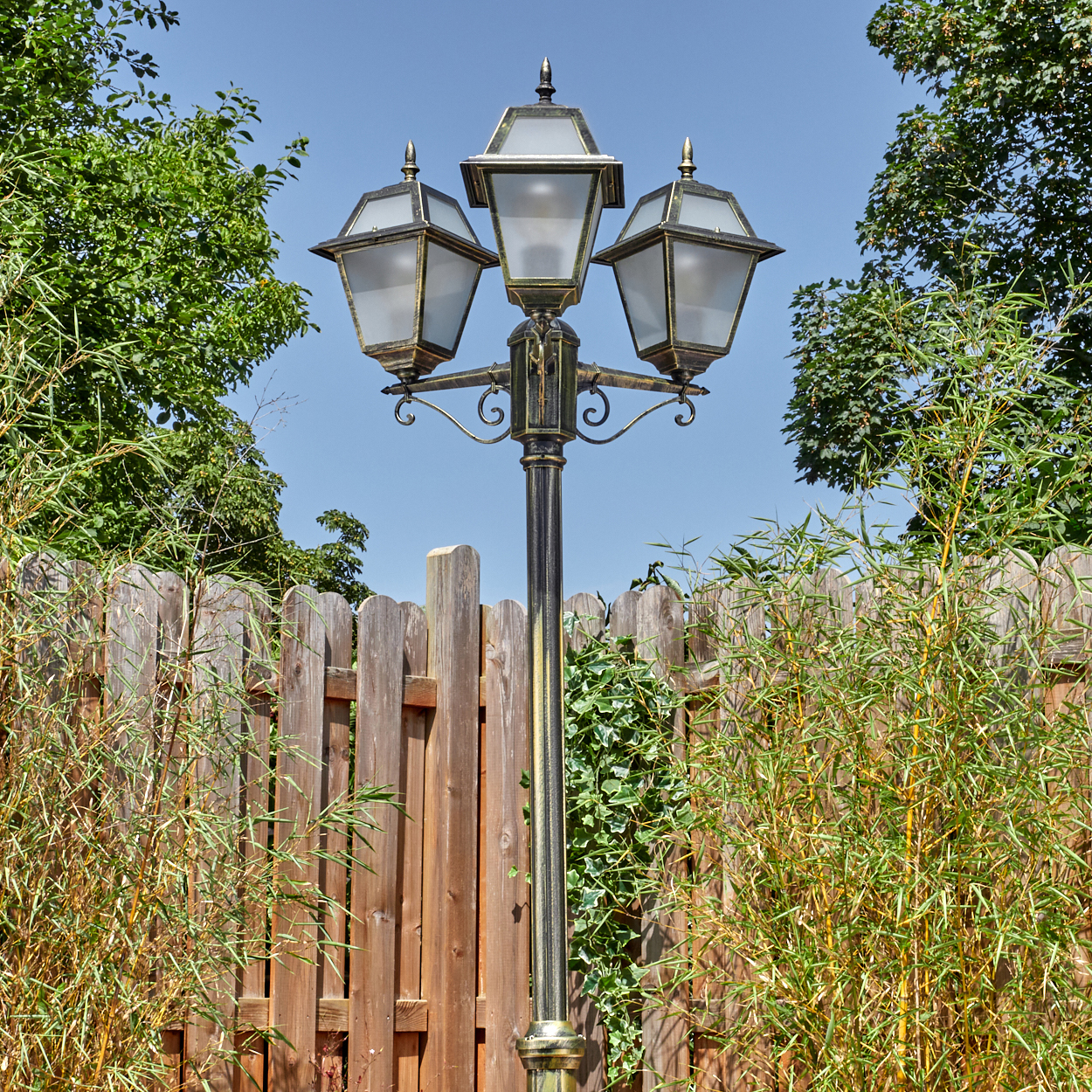 Eine dekorative Laterne mit drei Lampen steht vor einem Holzzaun. Grüne Bäume und Bambus umgeben sie unter blauem Himmel.