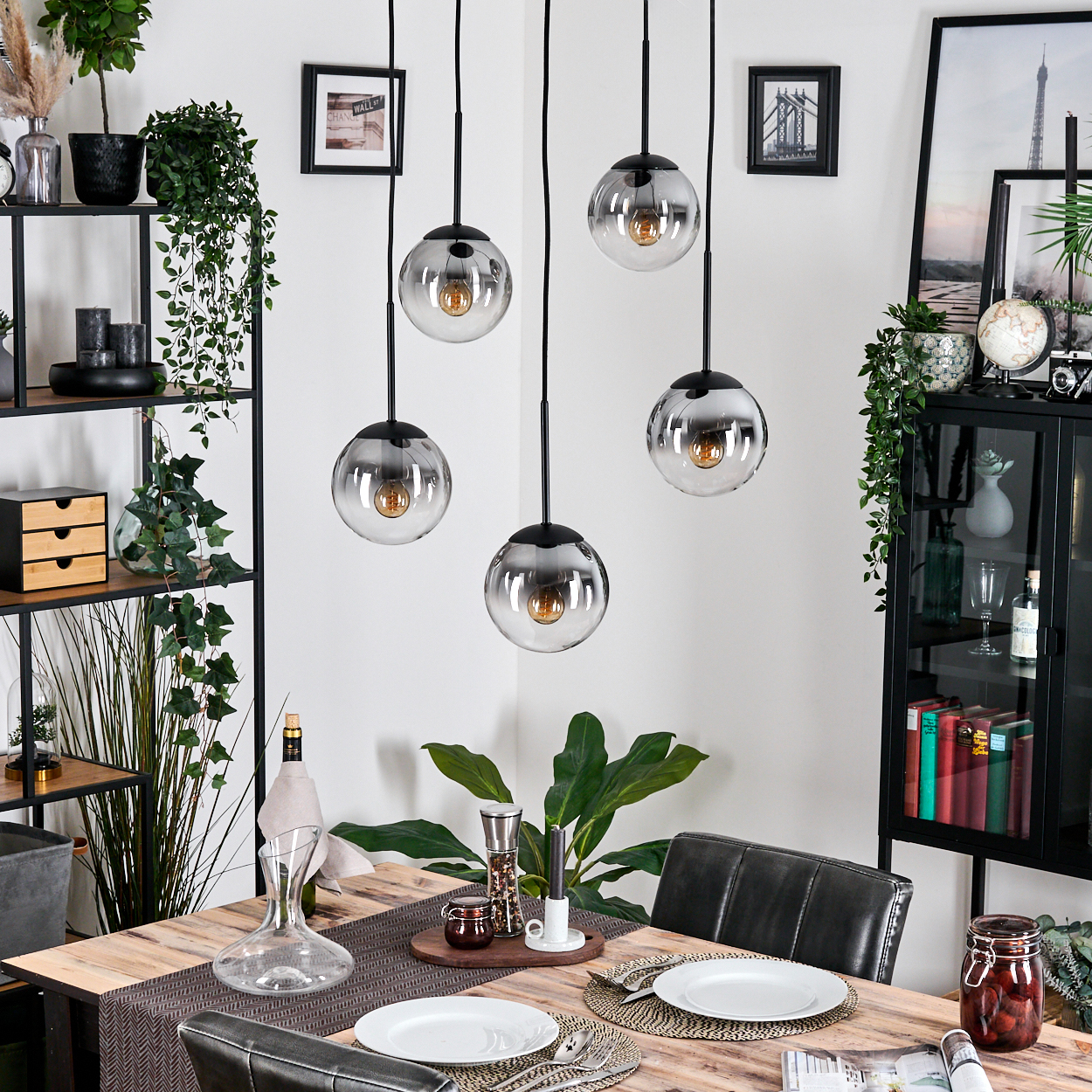 Esszimmer mit Holztisch, Hängelampen und Dekoration. Teller und Besteck. Pflanzen und ein Regal im Hintergrund.