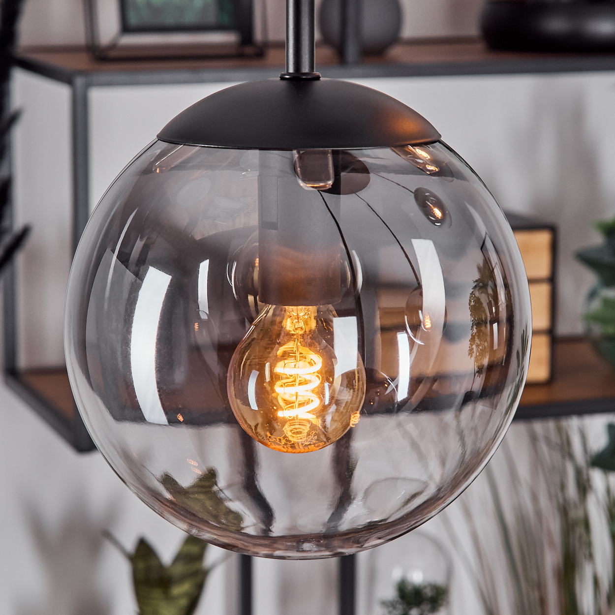 Eine moderne Pendelleuchte mit einer Glaskugel, die eine warme, glühende Glühbirne beherbergt. Schwarze Halterung, Regale im Hintergrund.