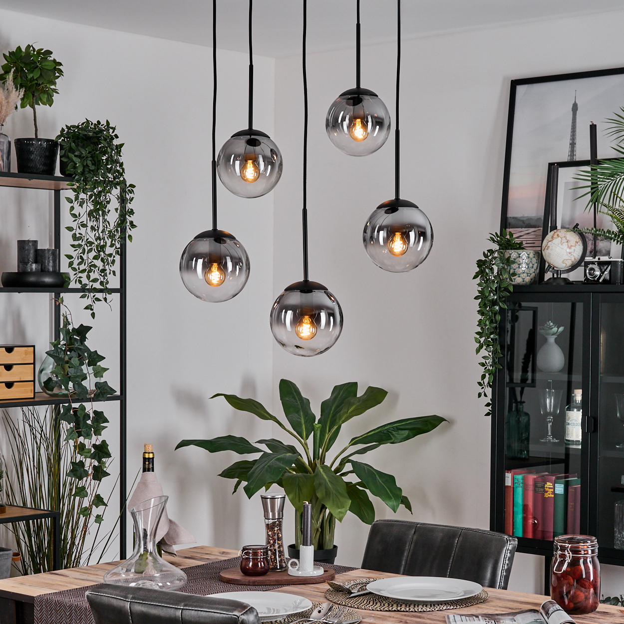 Esszimmer mit Holztisch, hängenden Kugelleuchten, Pflanzen und einem schwarzen Schrank mit Dekoration und einer Weinflasche.