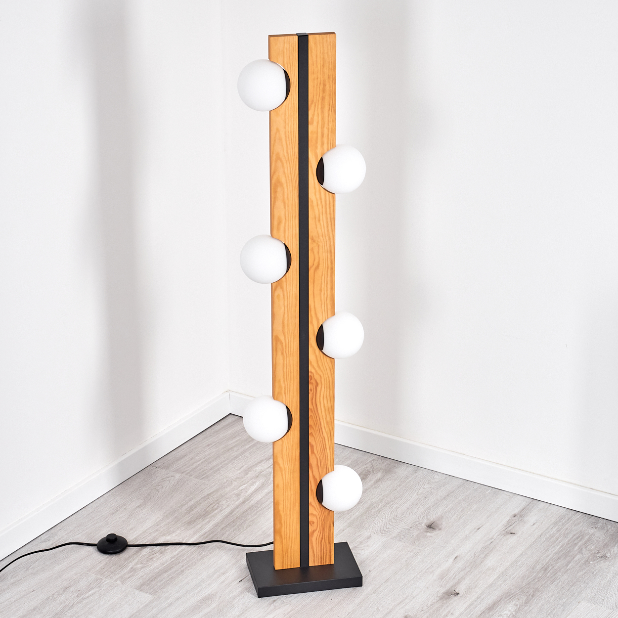 Eine moderne Stehlampe mit Holzrahmen und sechs weißen Kugelleuchten. Sie steht auf einem schwarzen Sockel. Der Hintergrund ist eine weiße Wand.