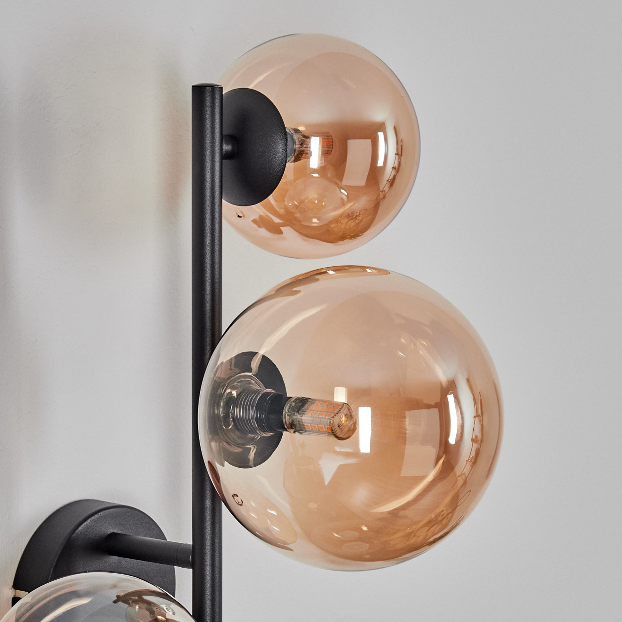 Eine moderne Wandlampe mit drei kugelförmigen, bernsteinfarbenen Glasschirmen und einem schwarzen Metallrahmen, an einer weißen Wand montiert.