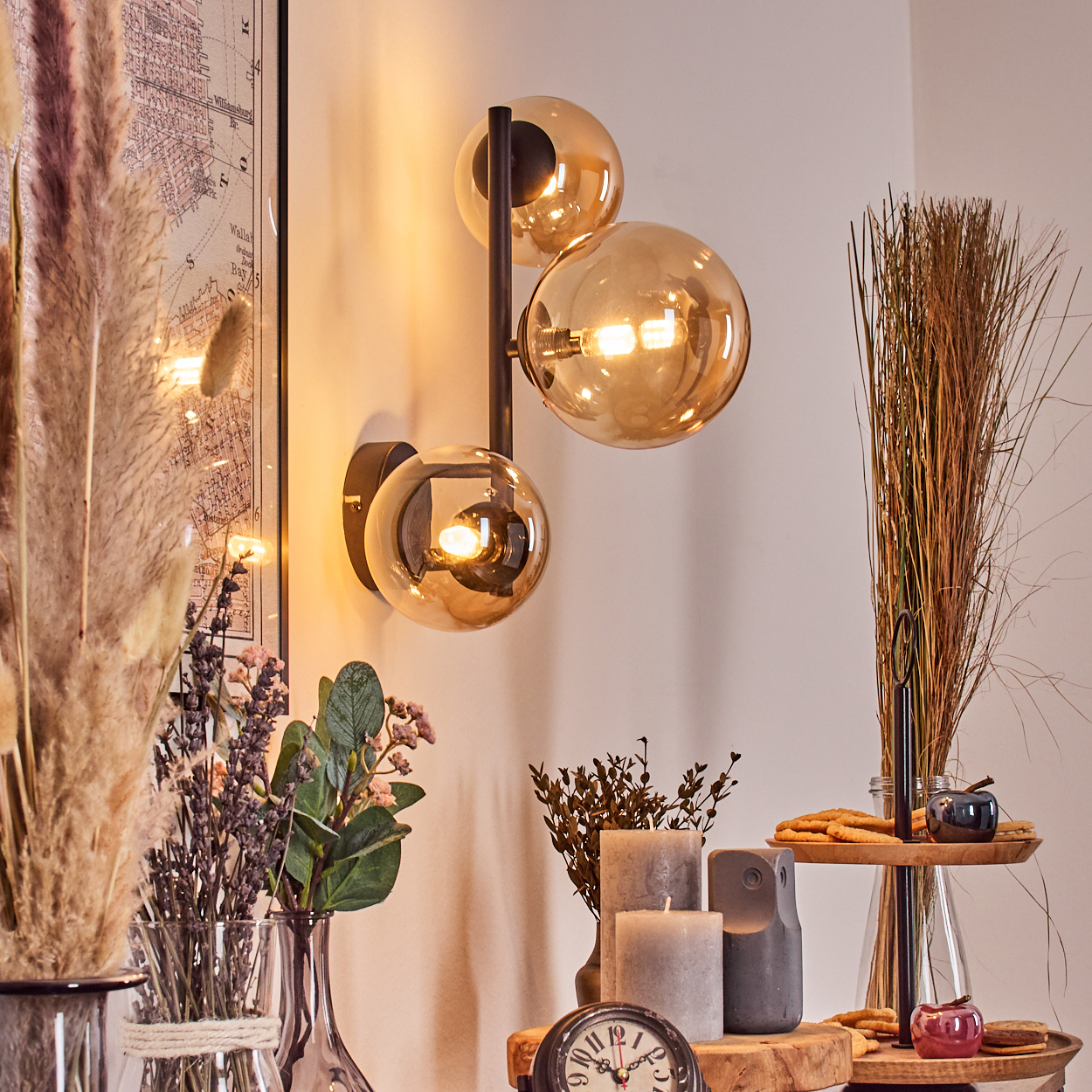 Eine Wandlampe mit drei Glaskugeln leuchtet. Getrocknete Pflanzen und Blumen stehen in Vasen. Kerzen, eine Uhr und Kekse stehen auf einem Holztisch.