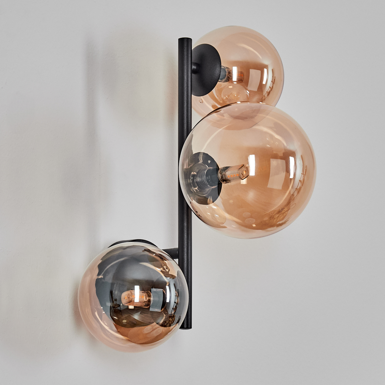 Eine Wandlampe mit drei kugelförmigen, bernsteinfarbenen Glasschirmen, die an einem schwarzen Metallrahmen an einer weißen Wand befestigt sind.