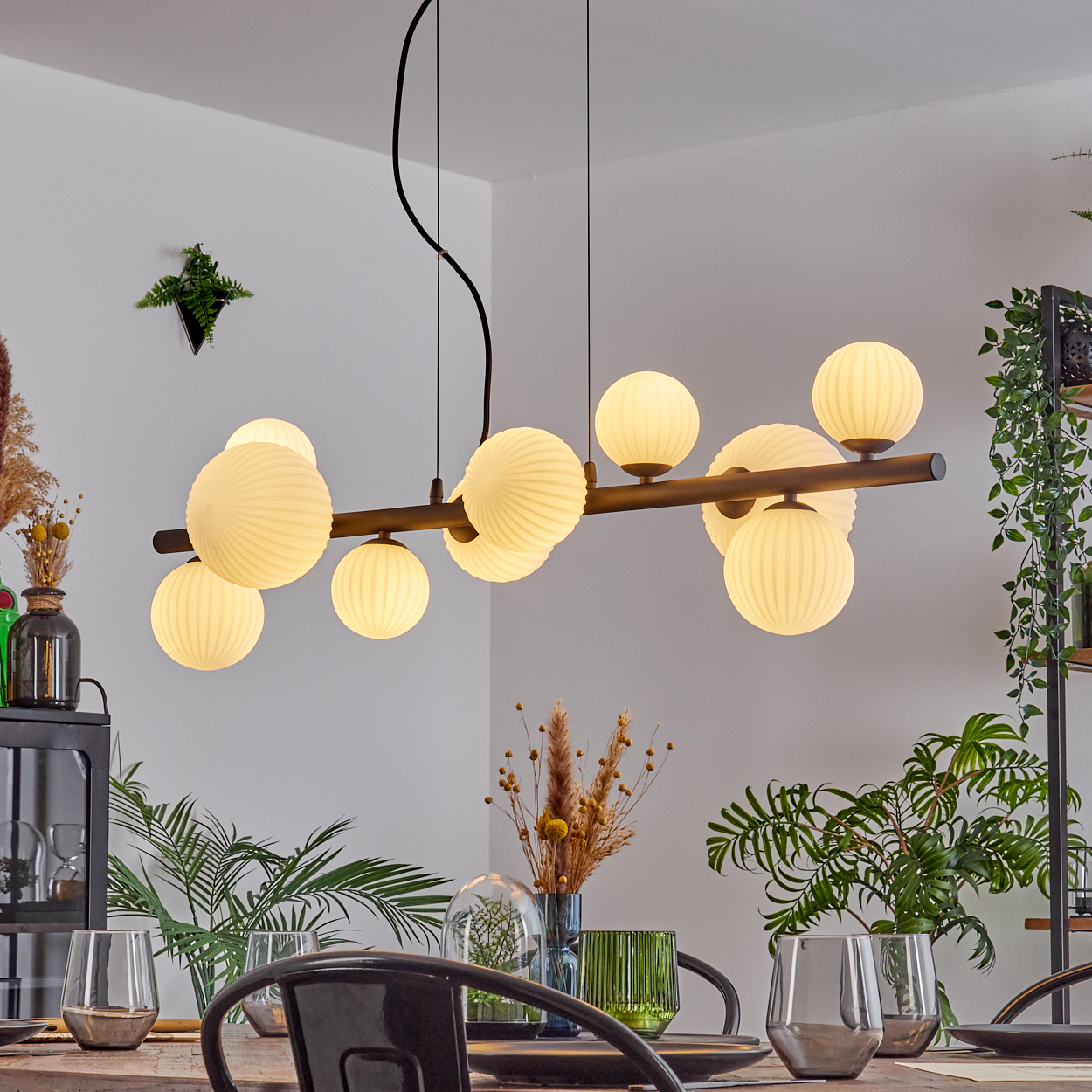 Ein Esszimmer mit einer Chandelier mit runden Lichtern. Pflanzen und Gläser auf dem Tisch. Weiße Wände, Holztisch.