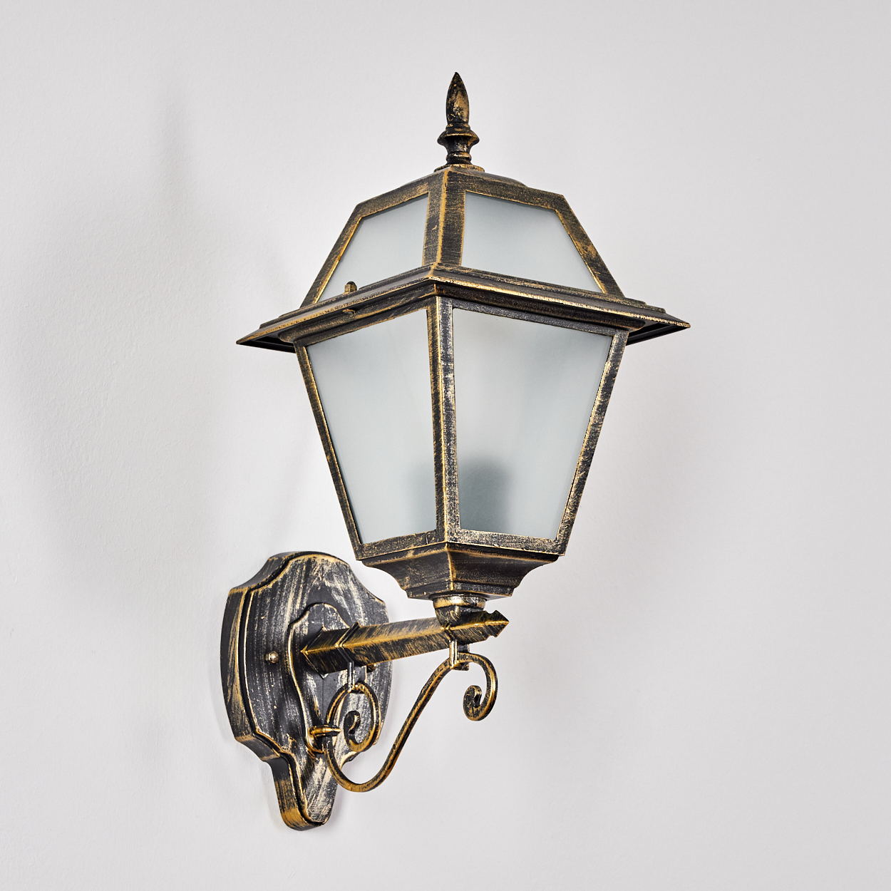 Eine verzierte Wandleuchte mit Bronze-Finish. Sie hat einen laternenförmigen Schirm mit Milchglasscheiben und sitzt an einer weißen Wand.