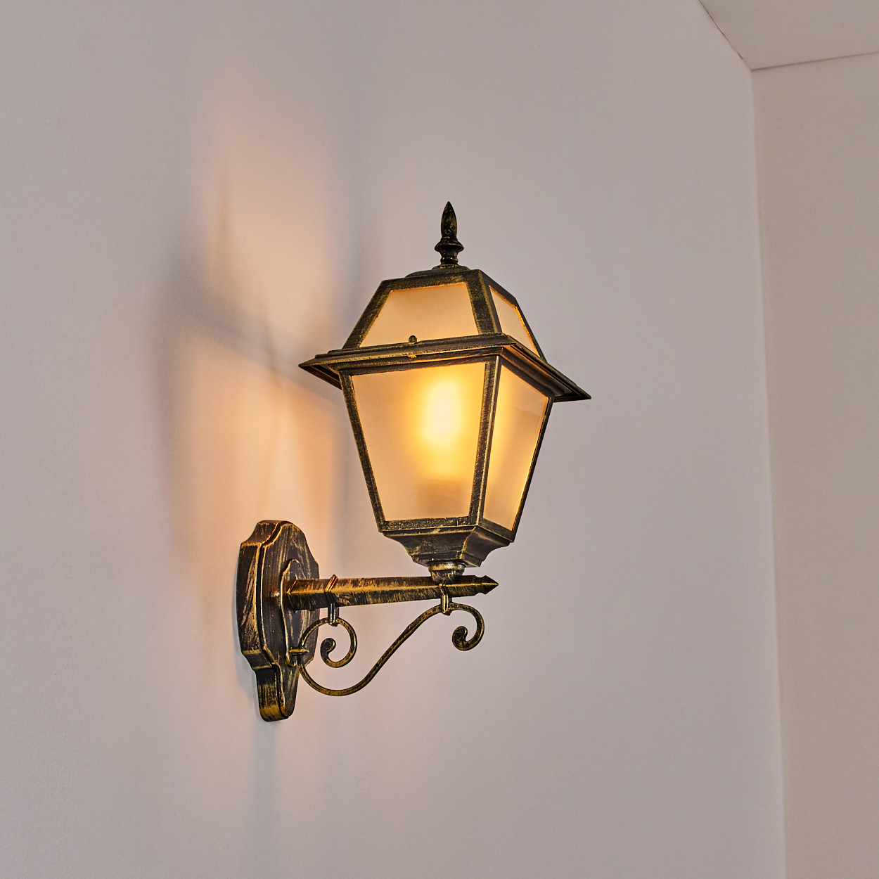 Eine Außenwandlaterne mit warmem Schein. Sie hat eine schwarz-goldene Oberfläche und ist an einer weißen Wand befestigt. Das Licht wirft einen Schatten.